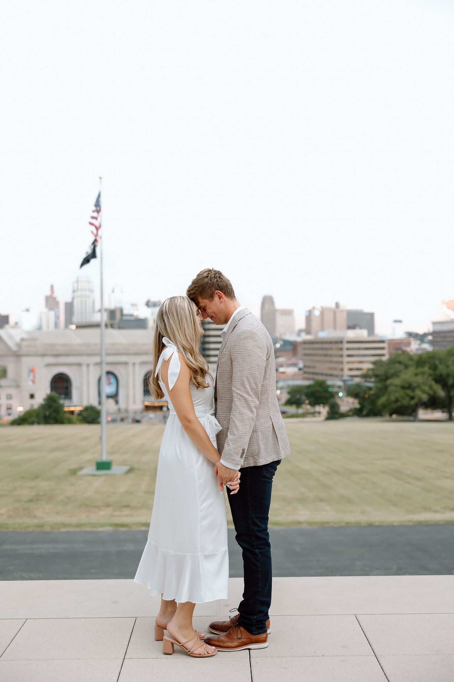 A couple shares a romantic moment on a rooftop overlooking a city skyline with green grass and buildings in view.