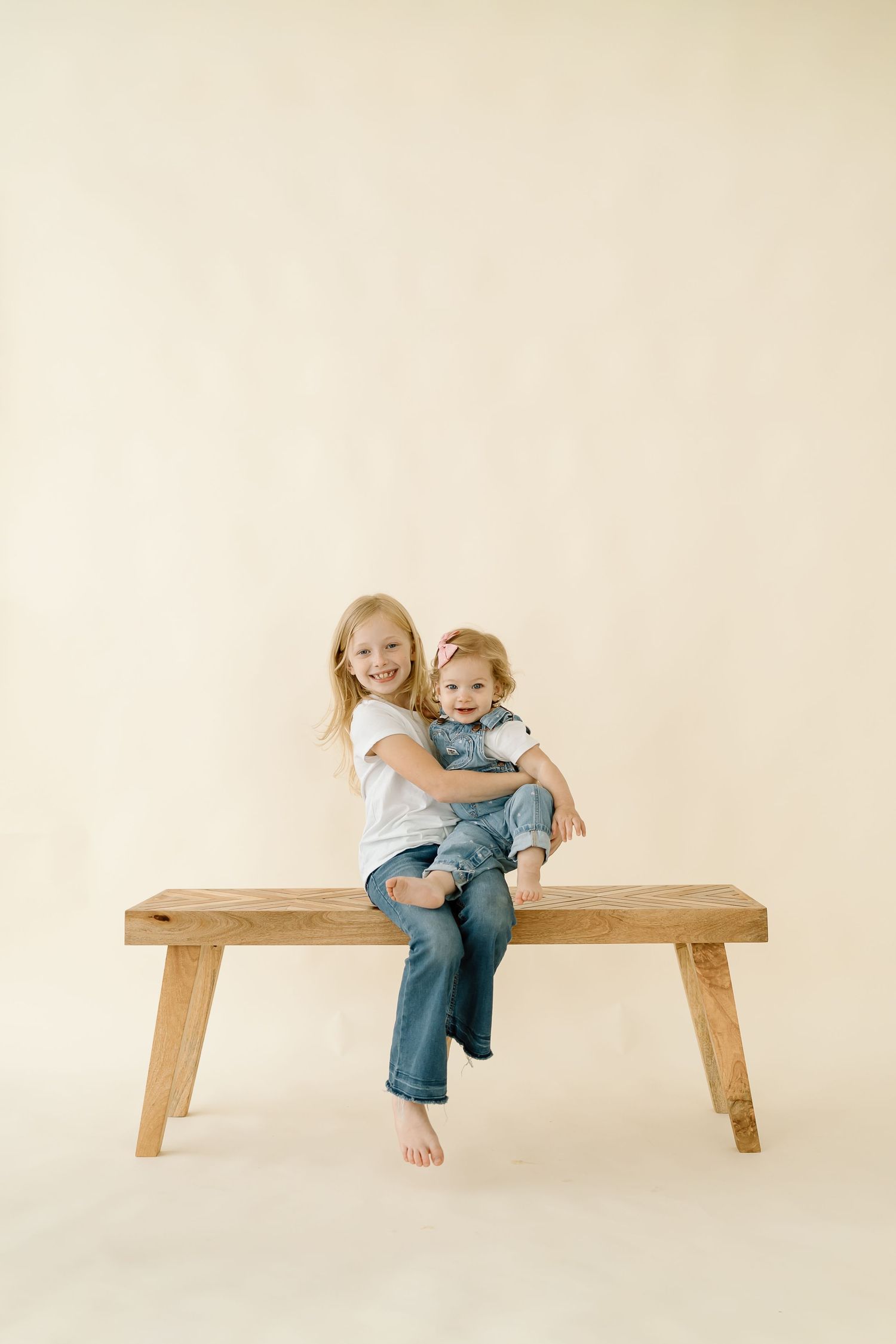 Two children sit together on a wooden bench against a neutral beige background.