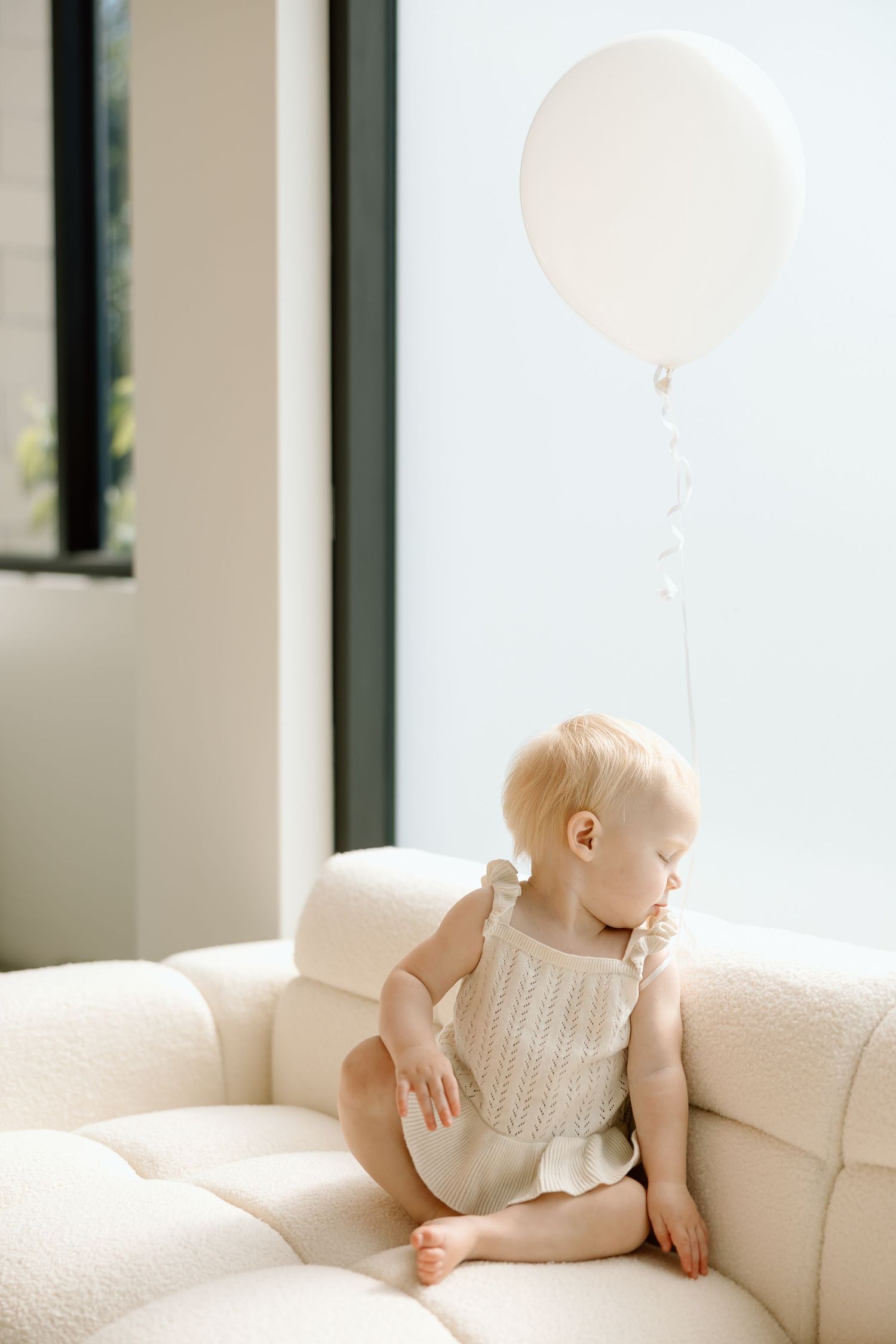 Baby in white romper crawls playfully on beige couch in sunlit room.