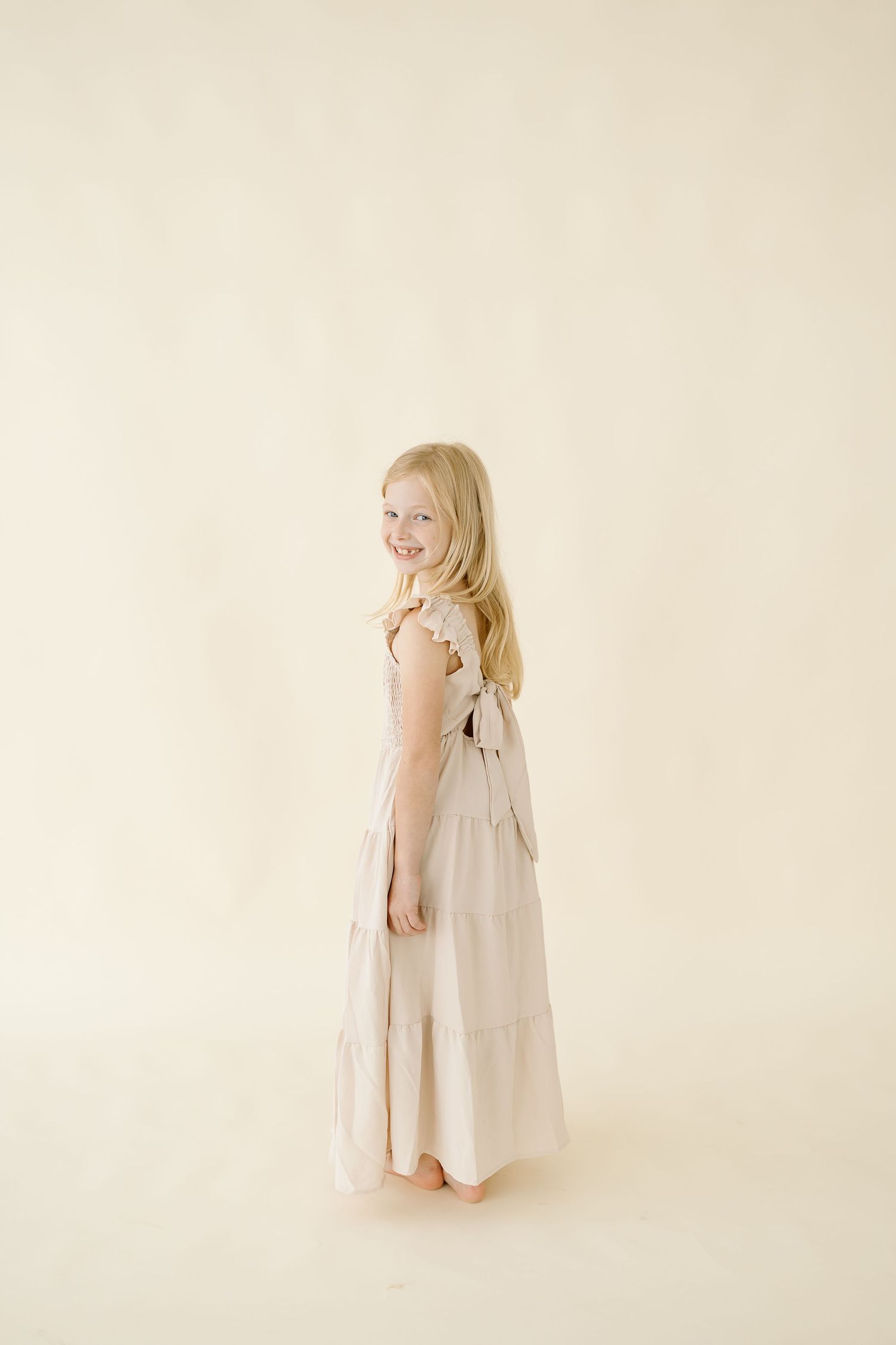 A young child stands sideways in a flowing beige maxi dress against a cream-colored backdrop.