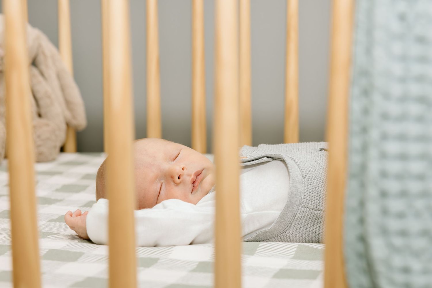 Baby sleeping peacefully in wooden crib with beige and gray bedding seen through crib slats.