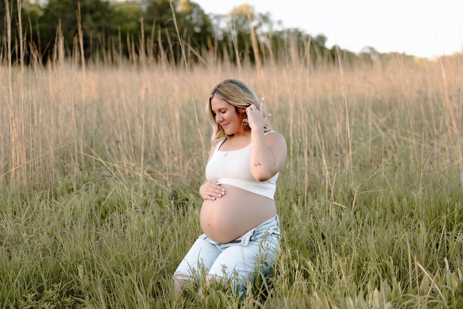 A pregnant woman in white sits peacefully in a golden field of tall grass during sunset.