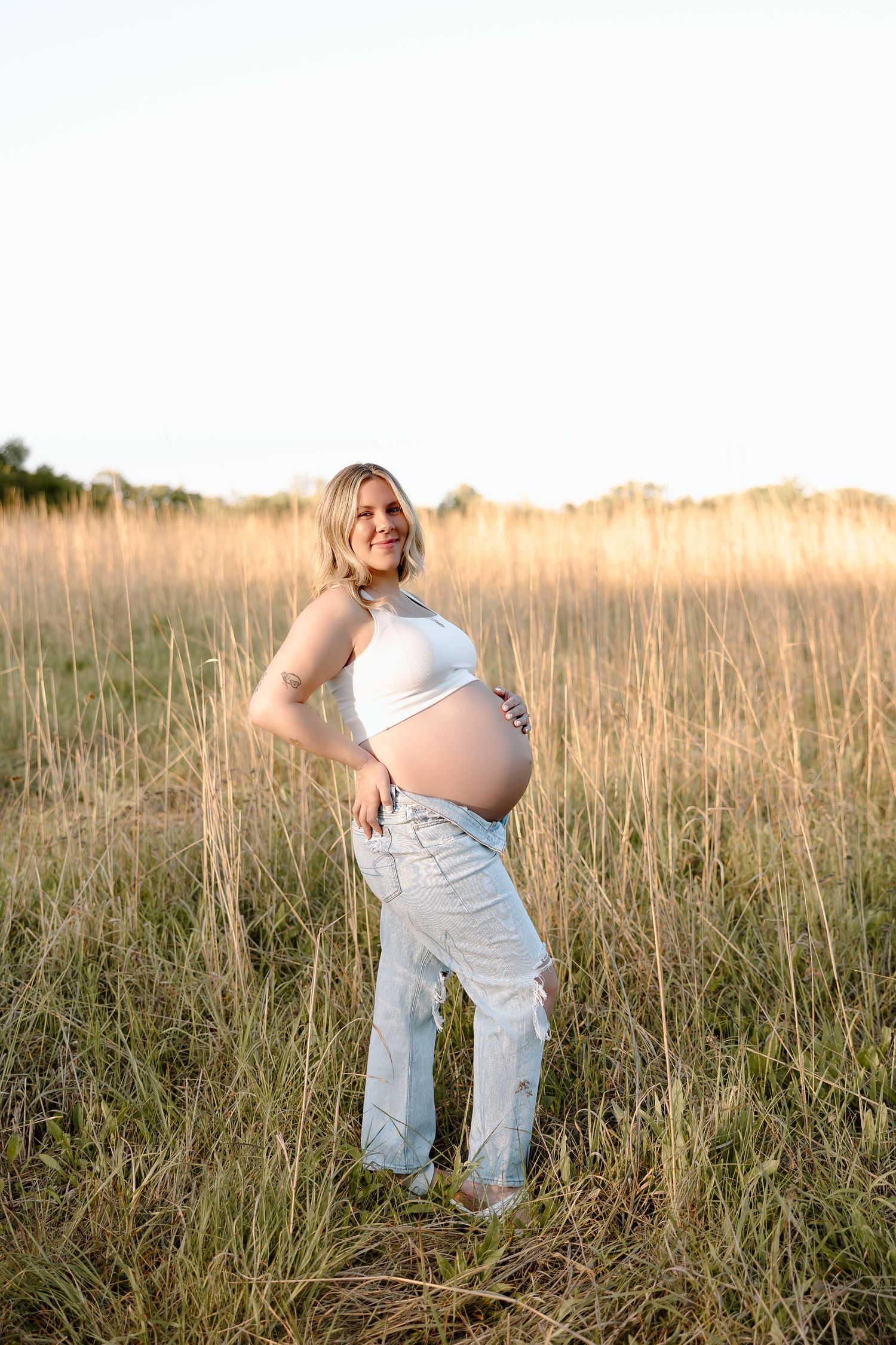 Person in white clothing poses expressively in a golden sunlit field of tall grass at sunset.