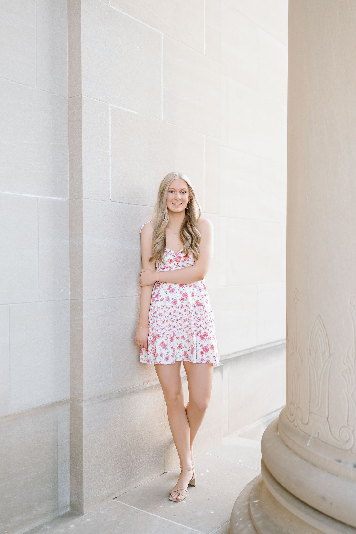A figure in a pink floral romper poses gracefully between white marble columns in a bright architectural setting.