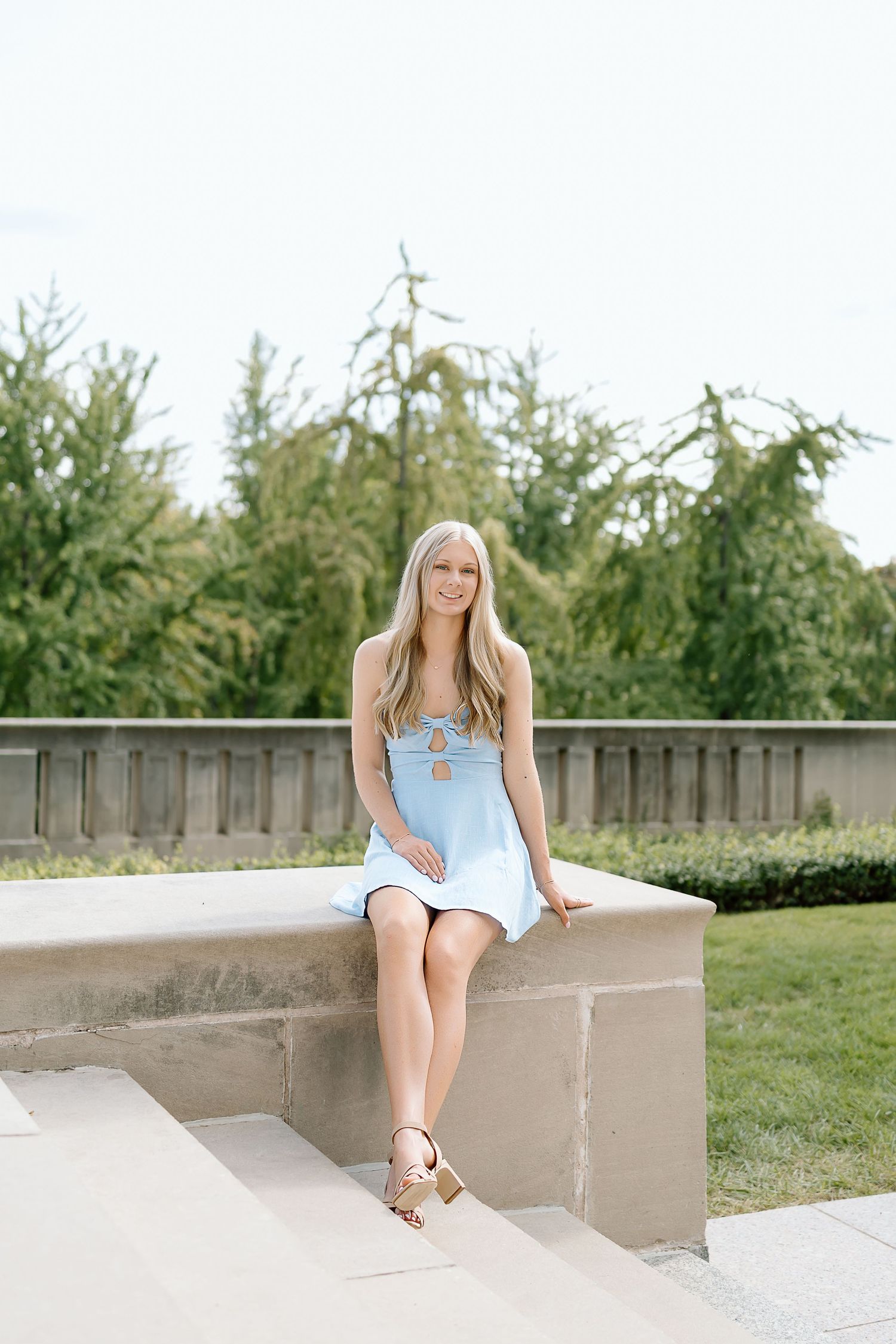 A woman in a light blue dress sits on a concrete ledge in a garden setting with evergreen trees in the background.