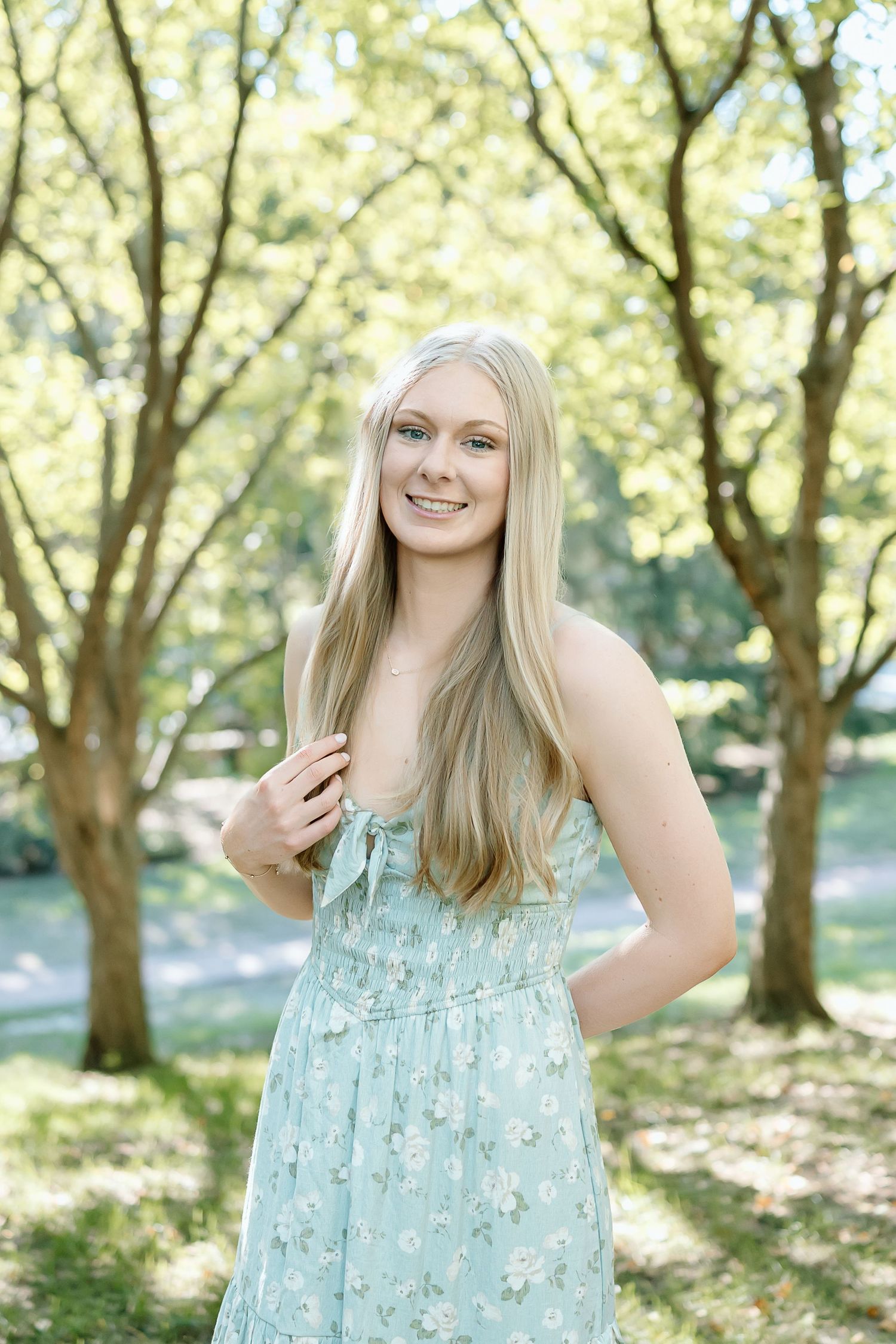 A series of outdoor portraits showing someone in a light blue floral sundress standing among sunlit trees in a park setting.