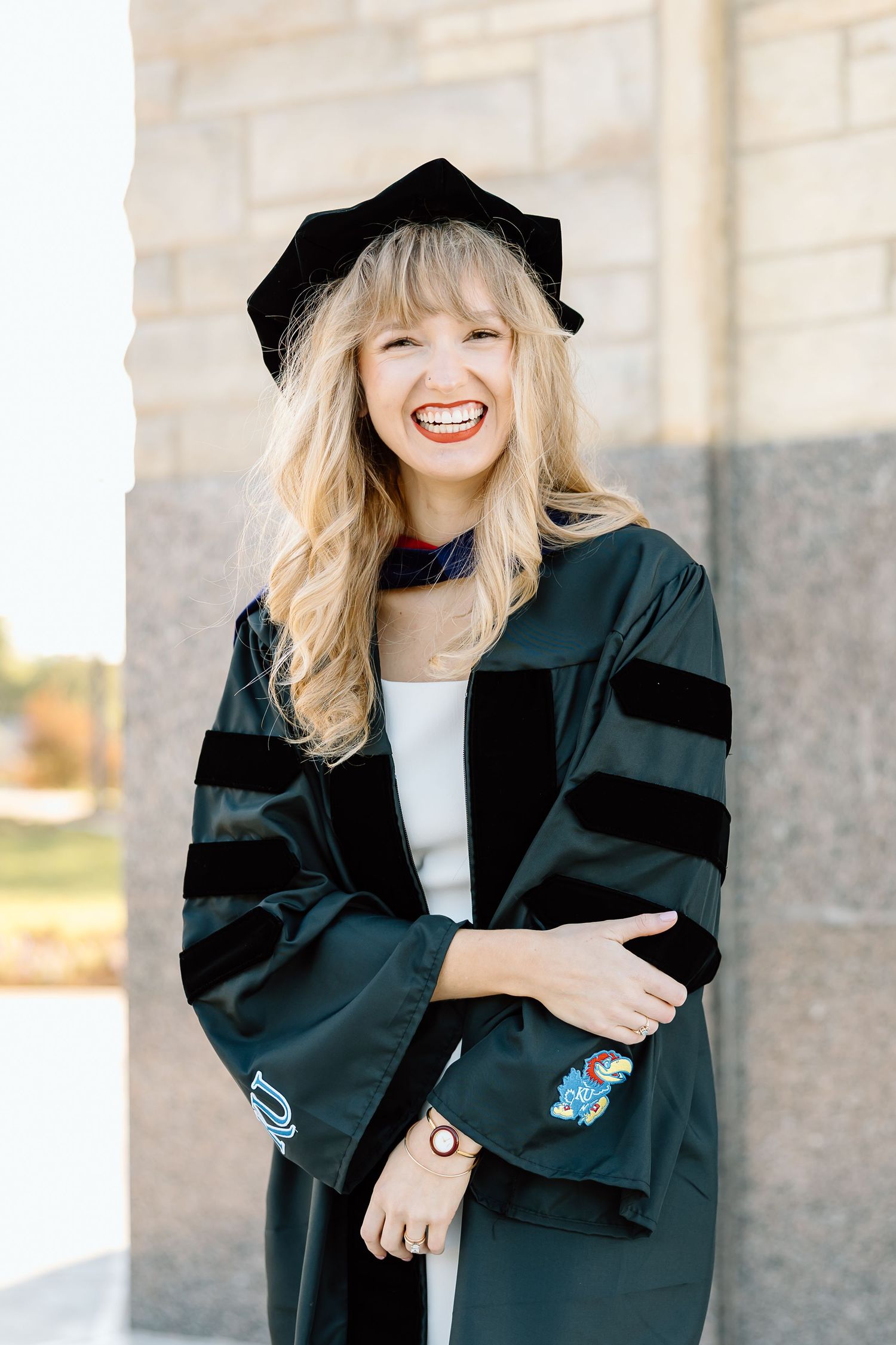 Graduate wearing traditional black academic regalia with velvet stripes and cap smiles joyfully during commencement ceremony.