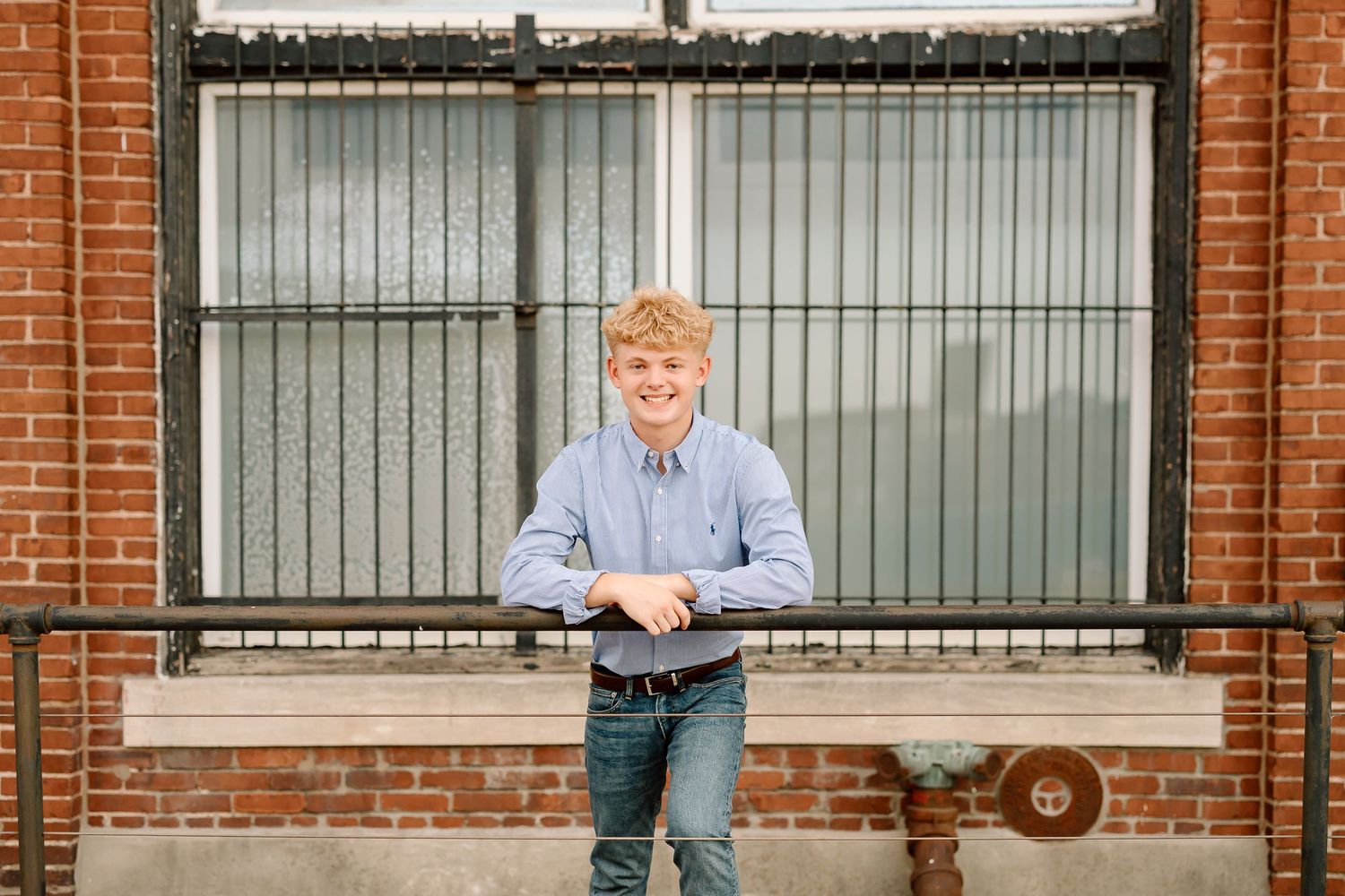 A person in a light blue button-down shirt stands smiling against a brick building exterior with black metal railings.