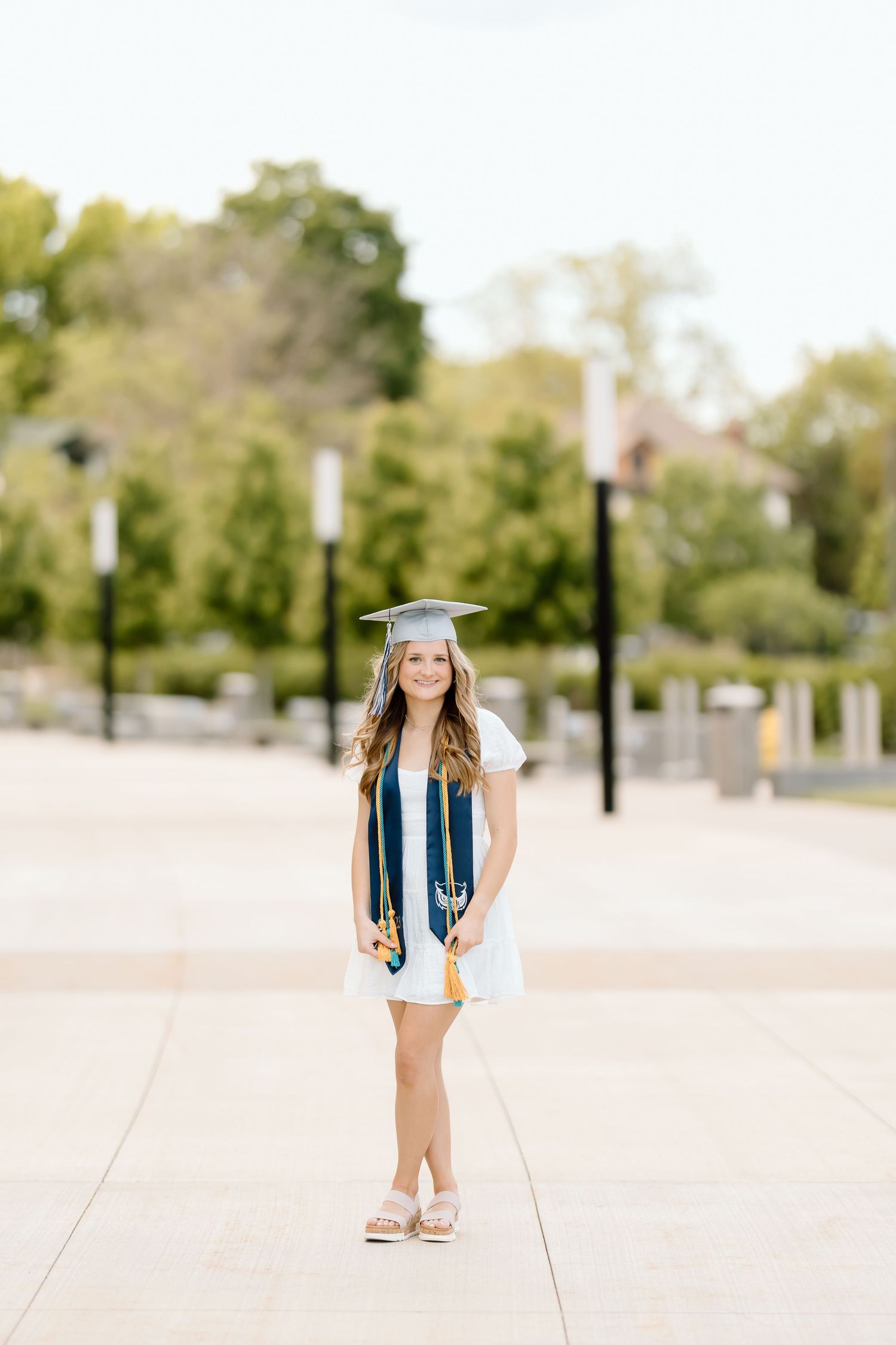 Graduate in blue cap and gown poses on outdoor plaza with lamp posts and trees.