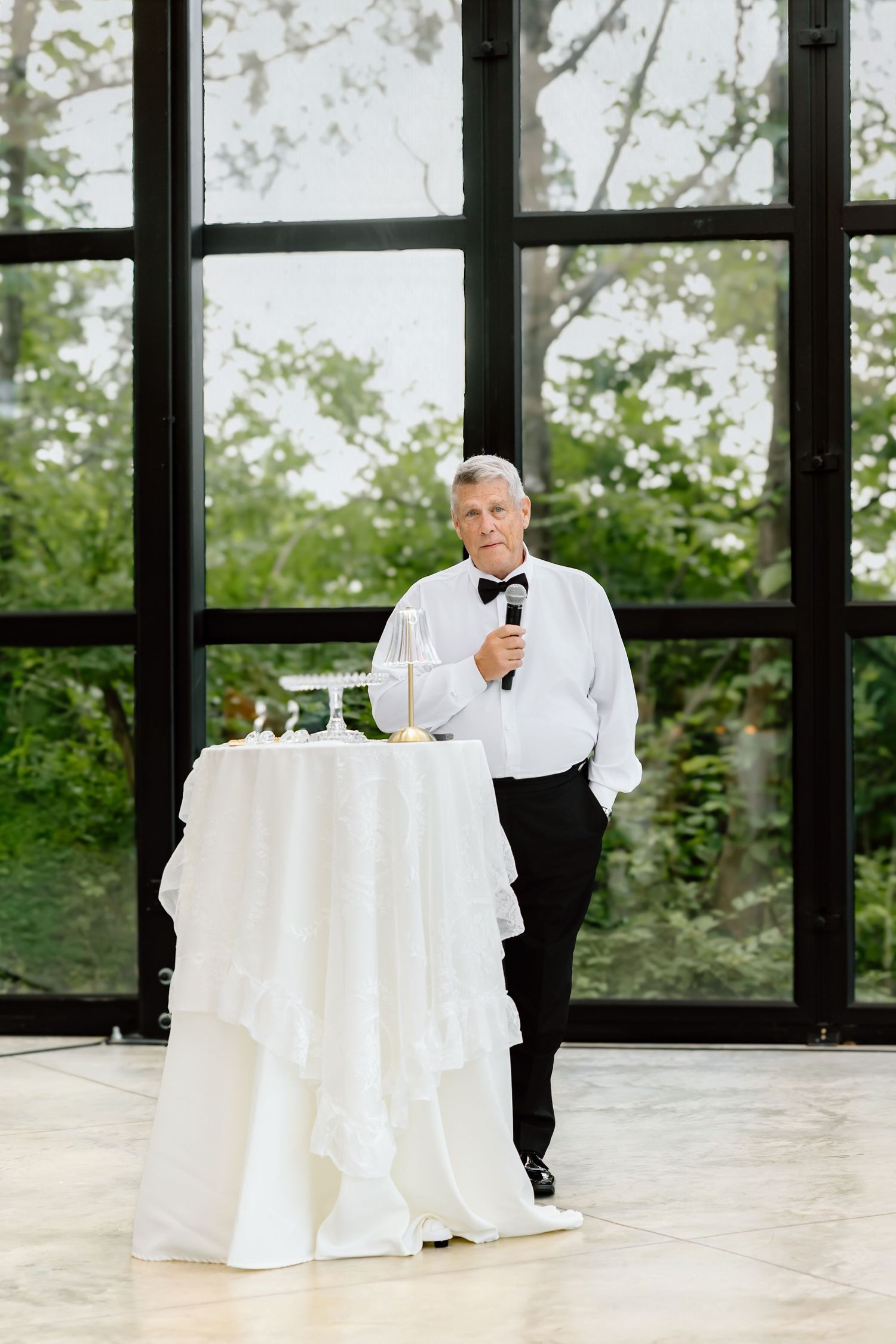 Elegant man in tuxedo holding microphone giving speech at wedding reception near white ruffled cocktail table.