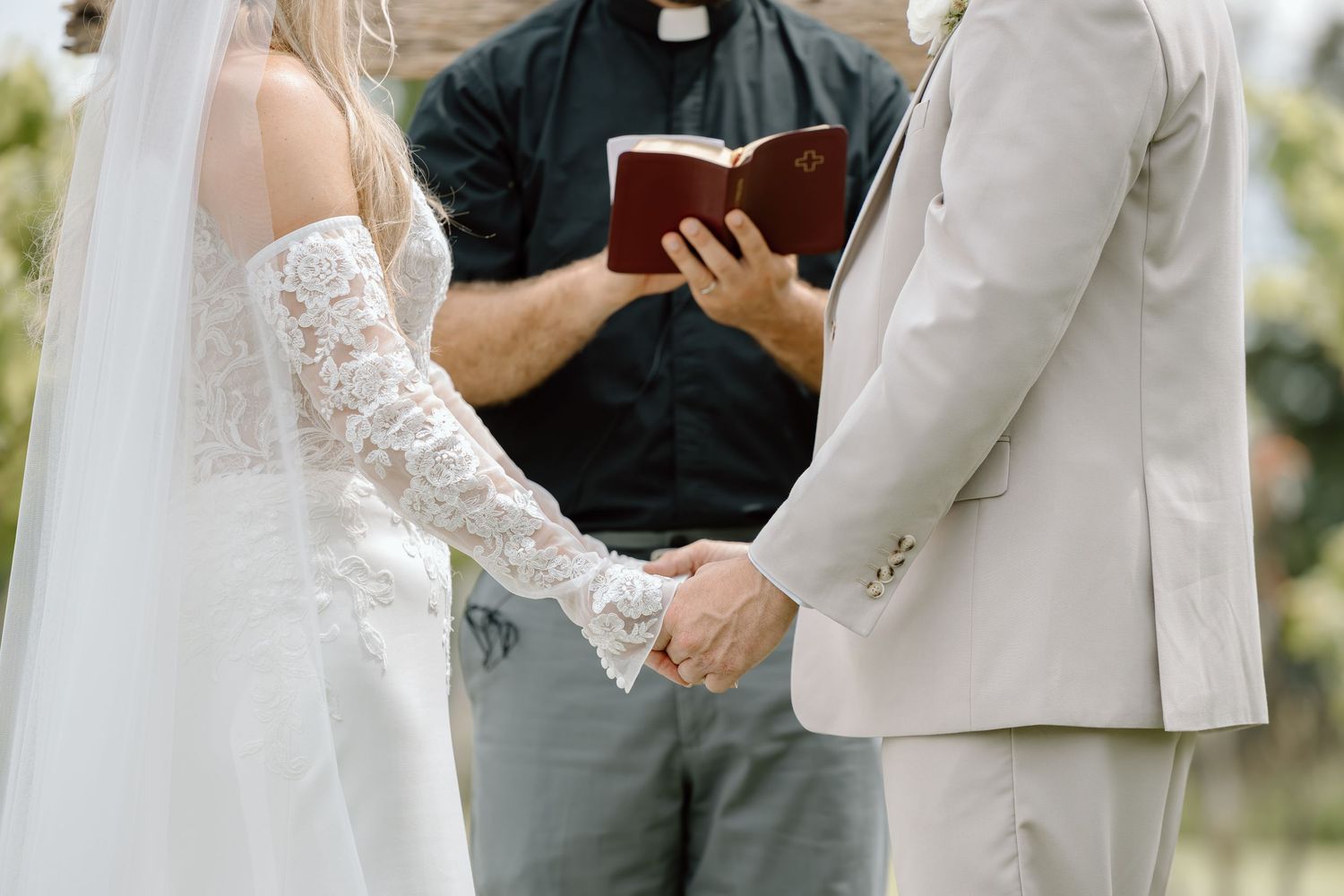 Bride and groom holding hands during outdoor wedding ceremony with officiant reading from book.