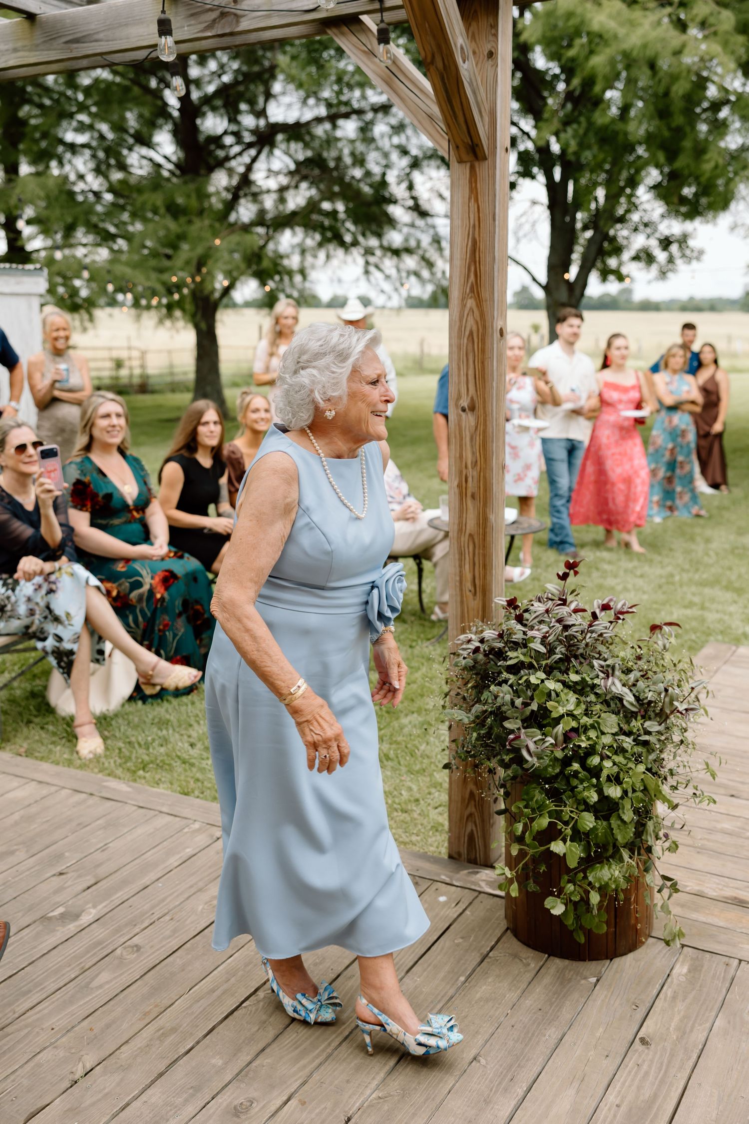 Elderly woman in light blue midi dress and floral shoes walks on wooden deck at outdoor wedding ceremony.