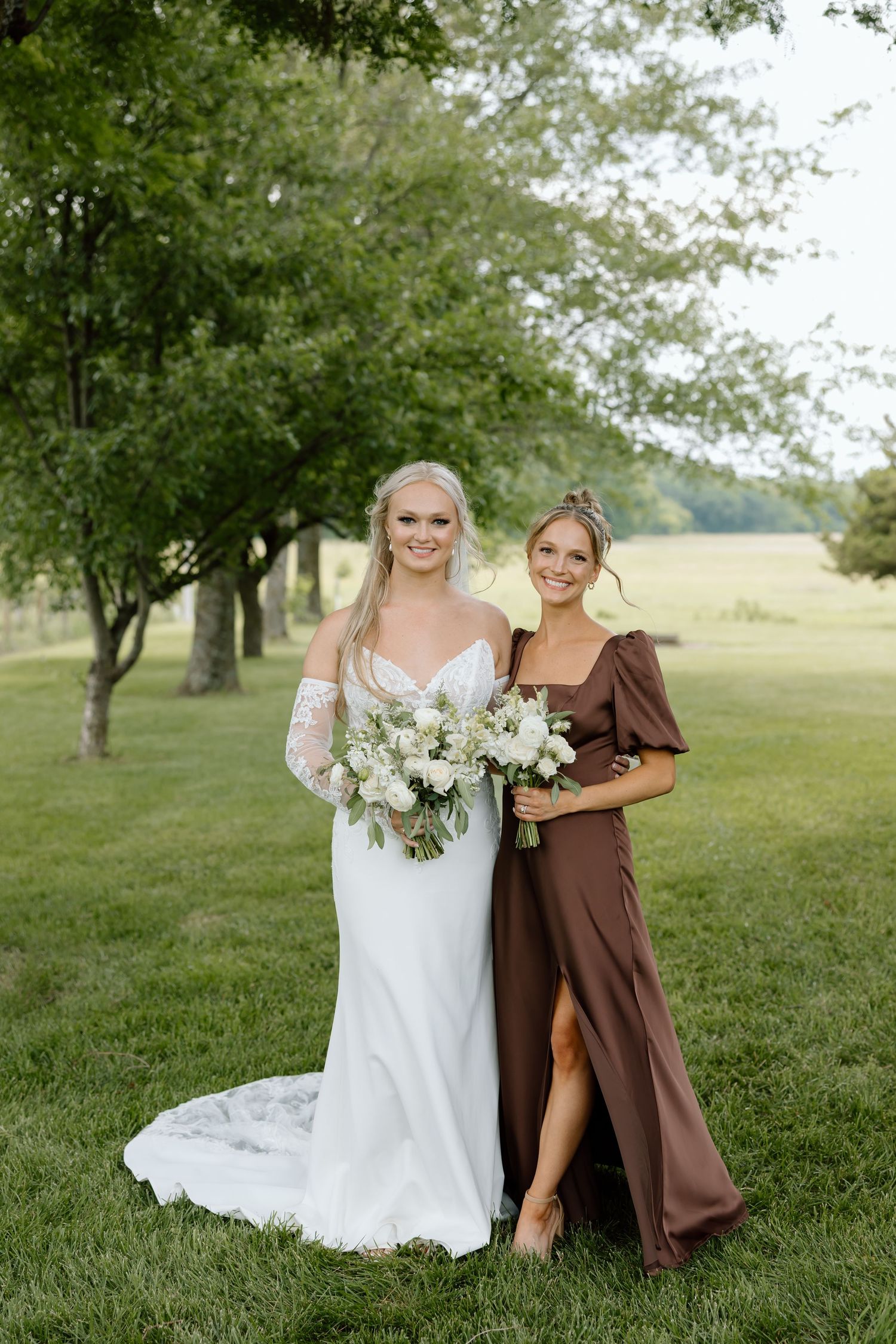 Bride in white gown with bouquet poses with bridesmaid in brown dress on green lawn.