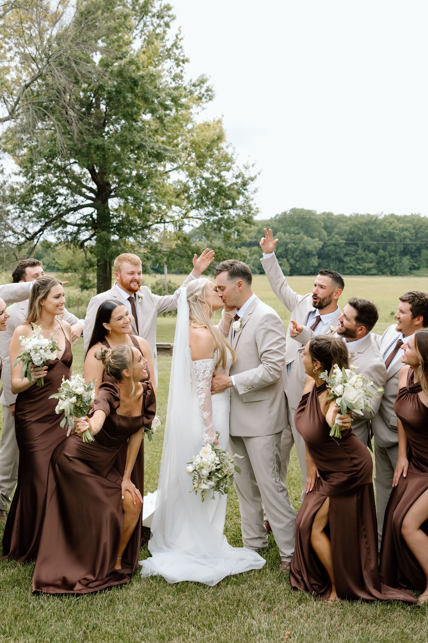 Wedding party celebrates as bride and groom kiss outdoors, bridesmaids in brown gowns and groomsmen in tan suits.