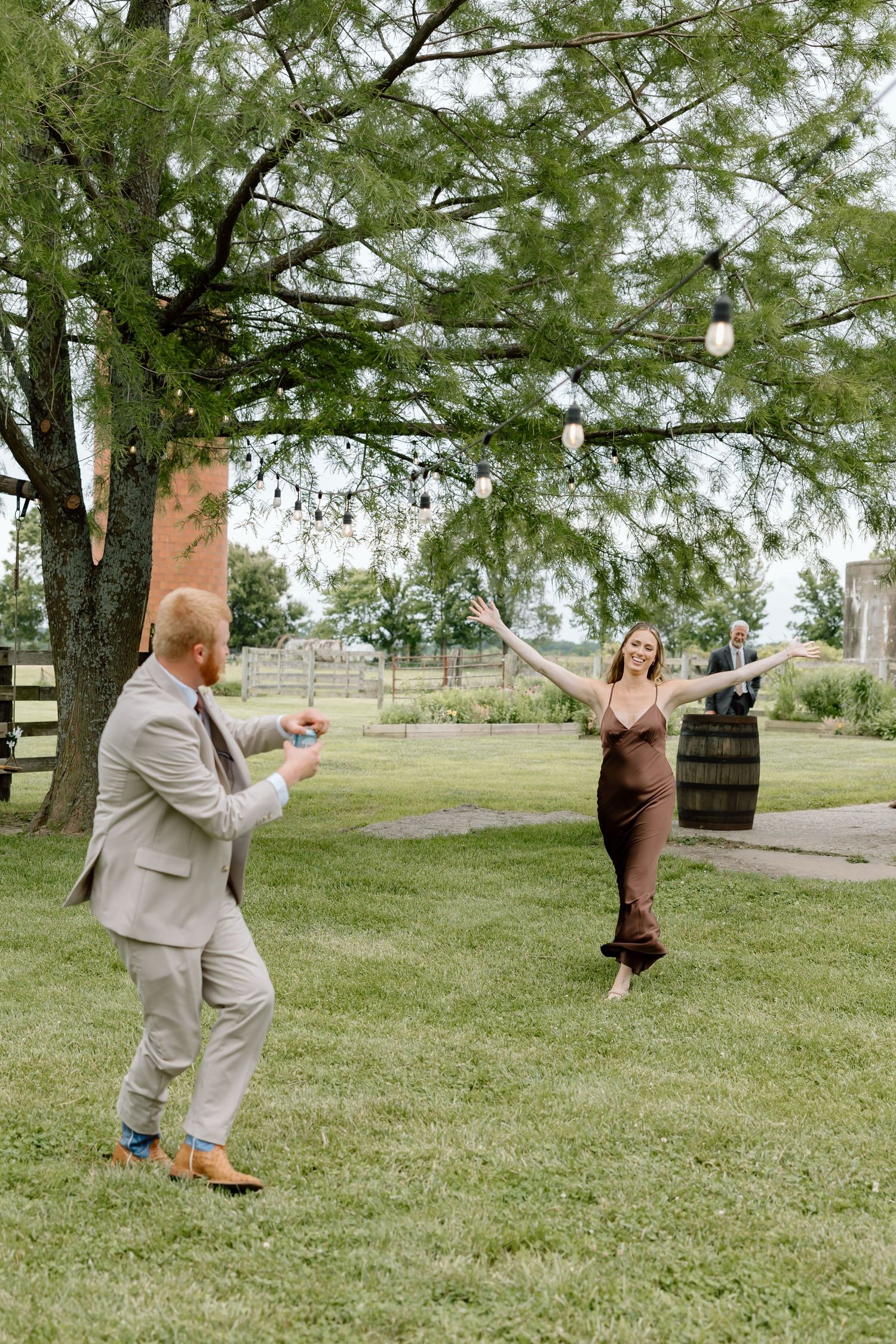 A man in a suit photographs a woman in a brown dress dancing outdoors at a rustic wedding venue.