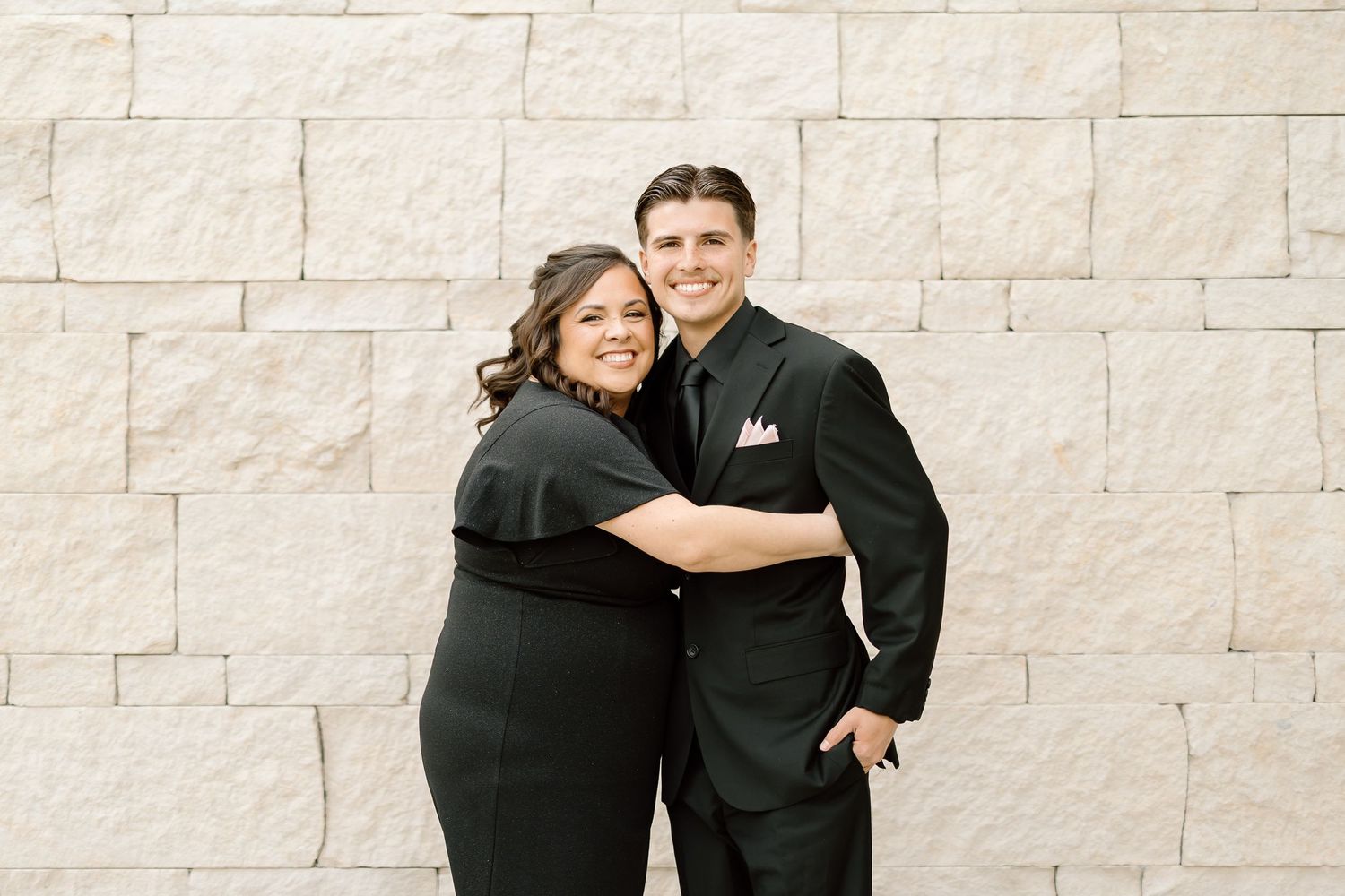 Couple in formal black attire embracing against a light stone wall background.