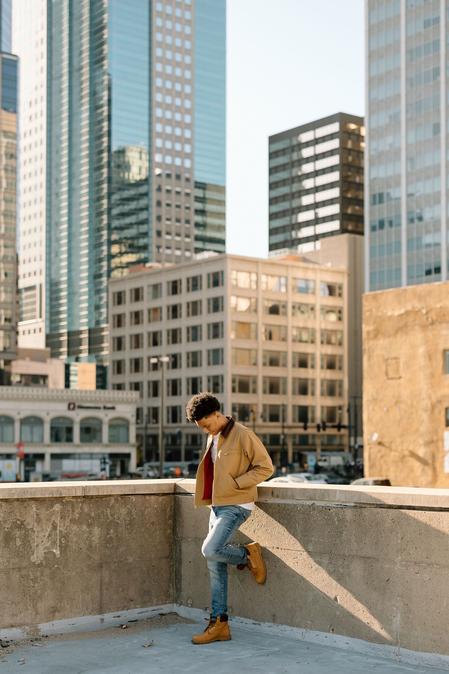 Person in beige jacket and jeans poses against cityscape on concrete rooftop.