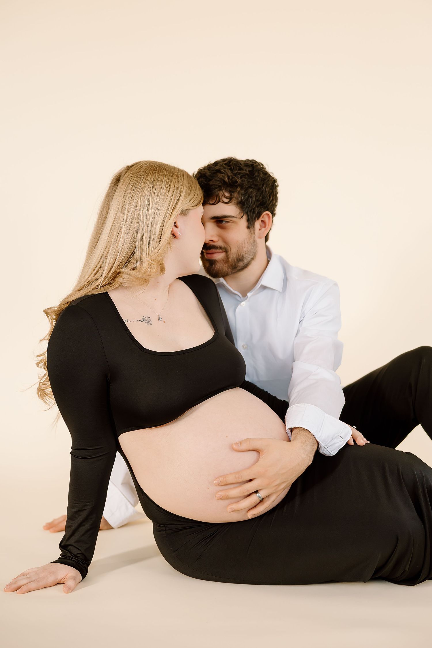 Tender moment between expectant couple during maternity photoshoot on neutral backdrop.