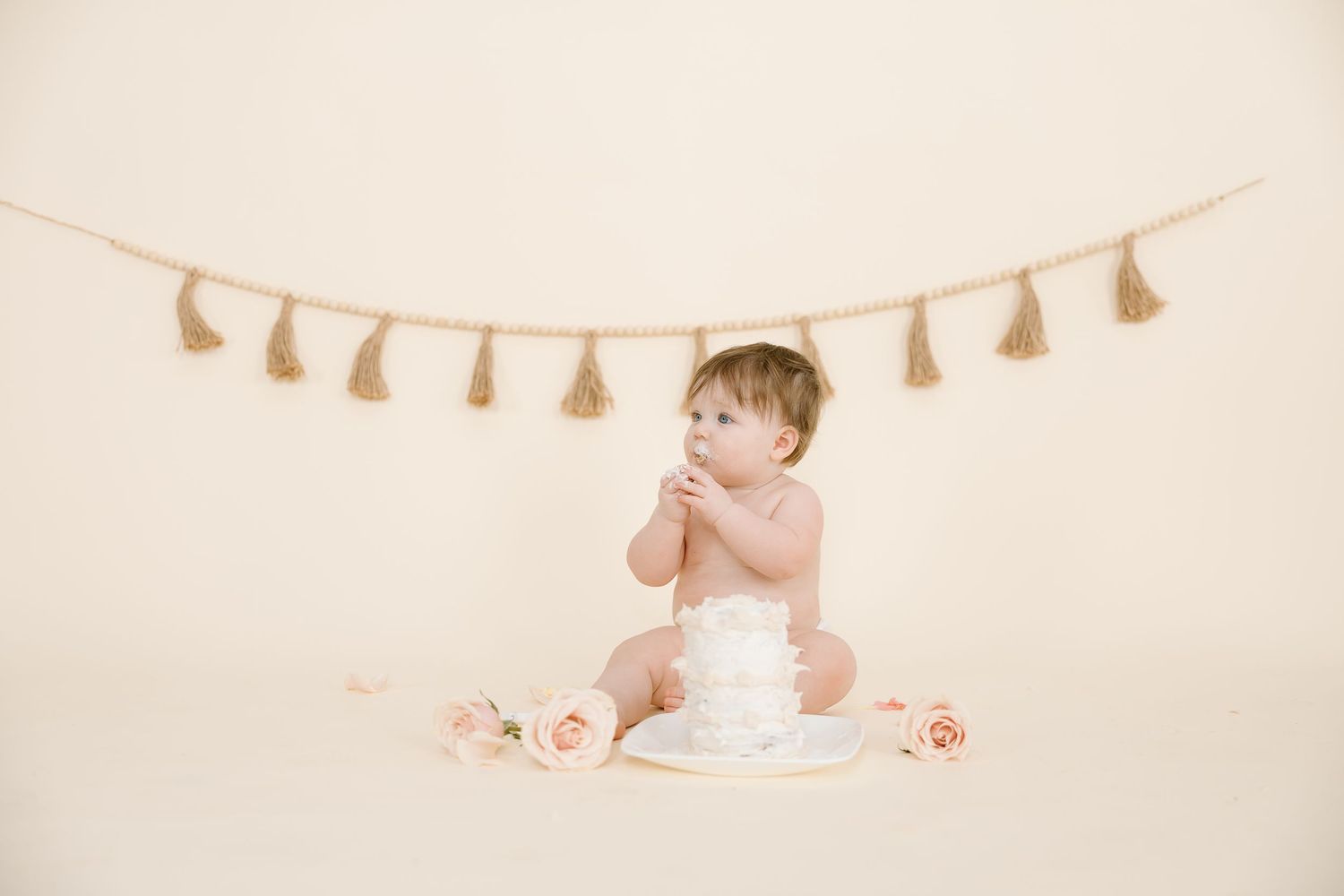 A baby poses with hands near face beside white cake under decorative tassel garland.
