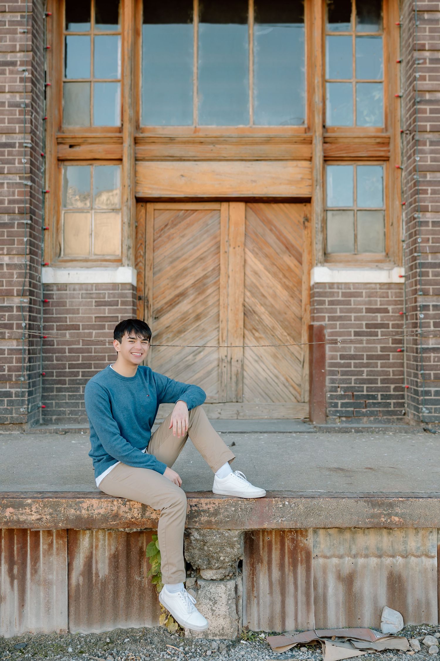 Casual portrait of someone in blue sweater sitting on concrete ledge against rustic wooden door and brick wall.