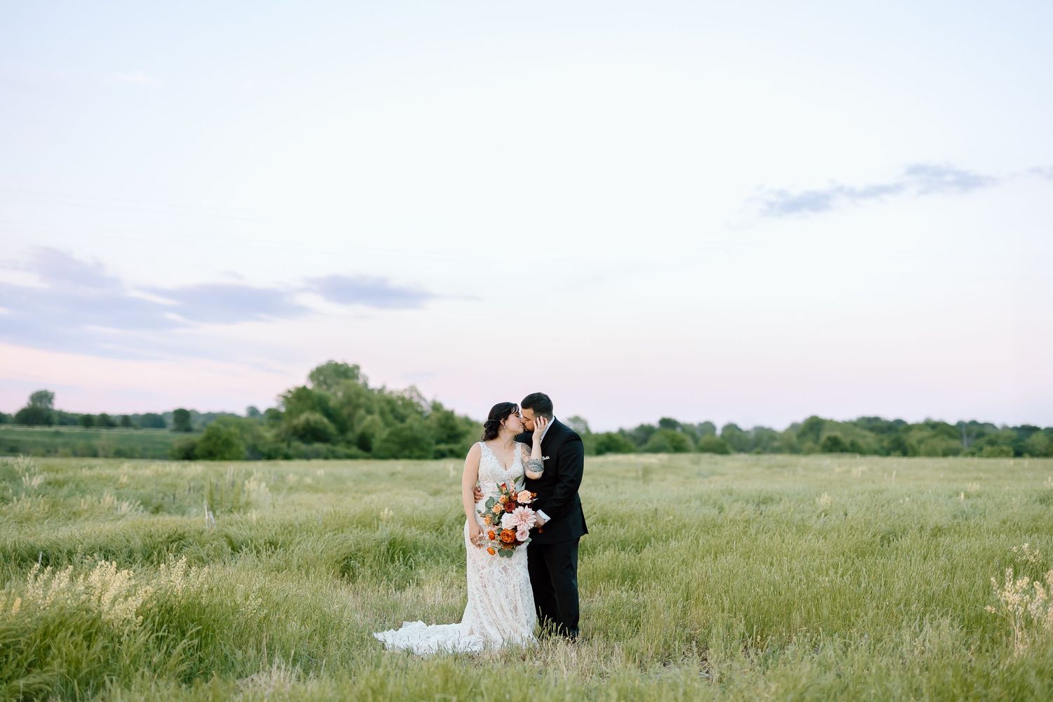 Couple in wedding attire embraces in a sun-drenched meadow with tall prairie grass stretching to the horizon.
