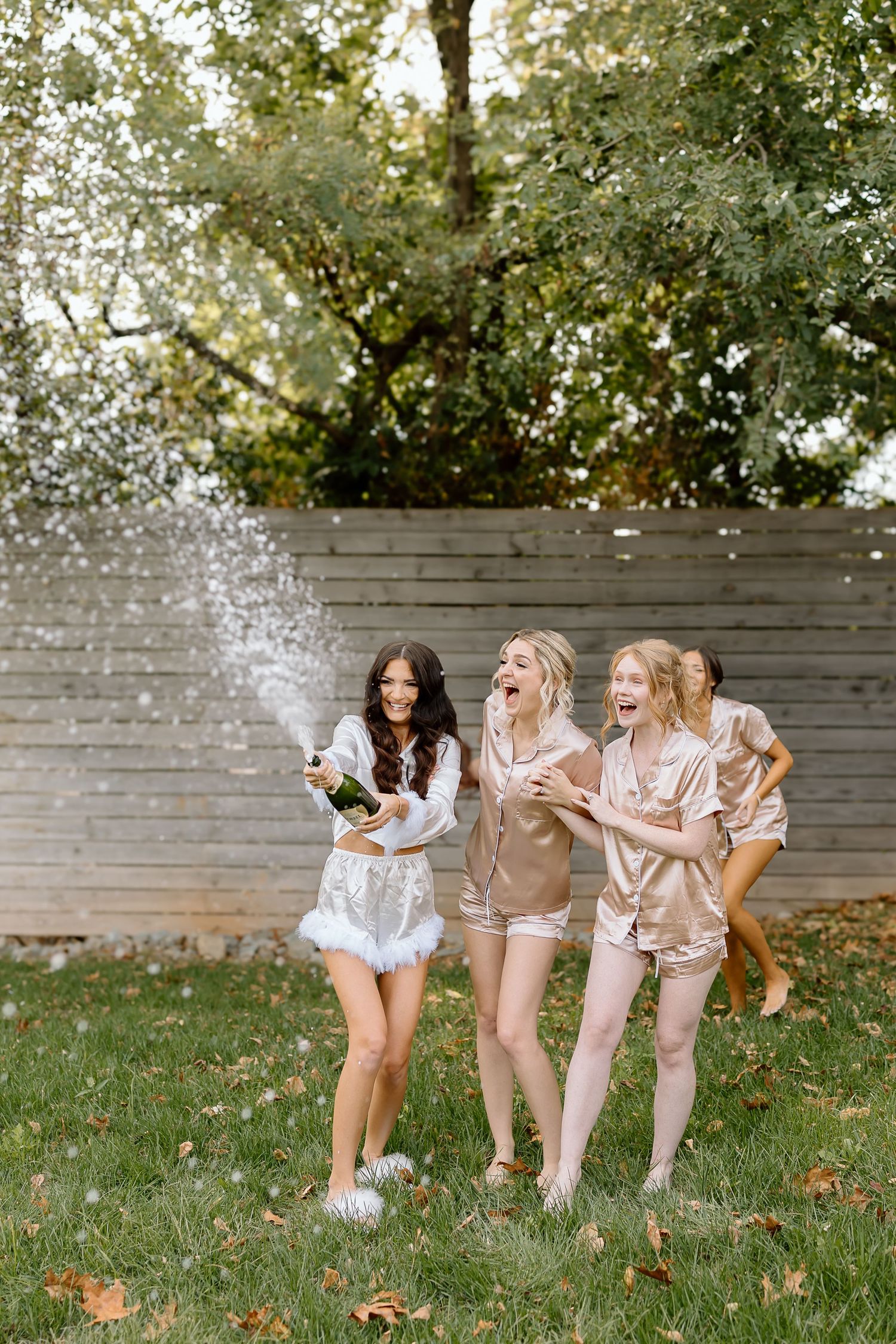 Three friends in casual outfits spraying champagne and celebrating together on a grassy lawn.
