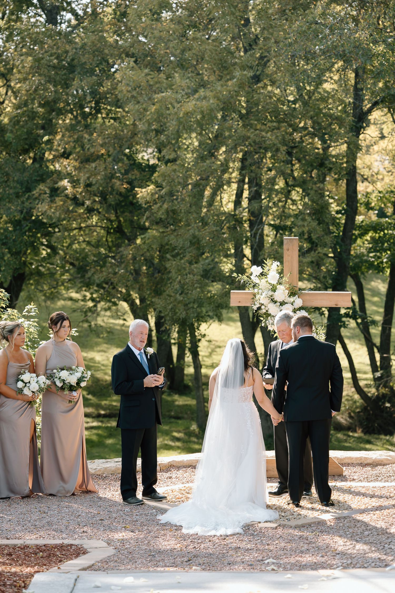 Wedding party poses for formal outdoor photos in a wooded area with gravel path and natural lighting.