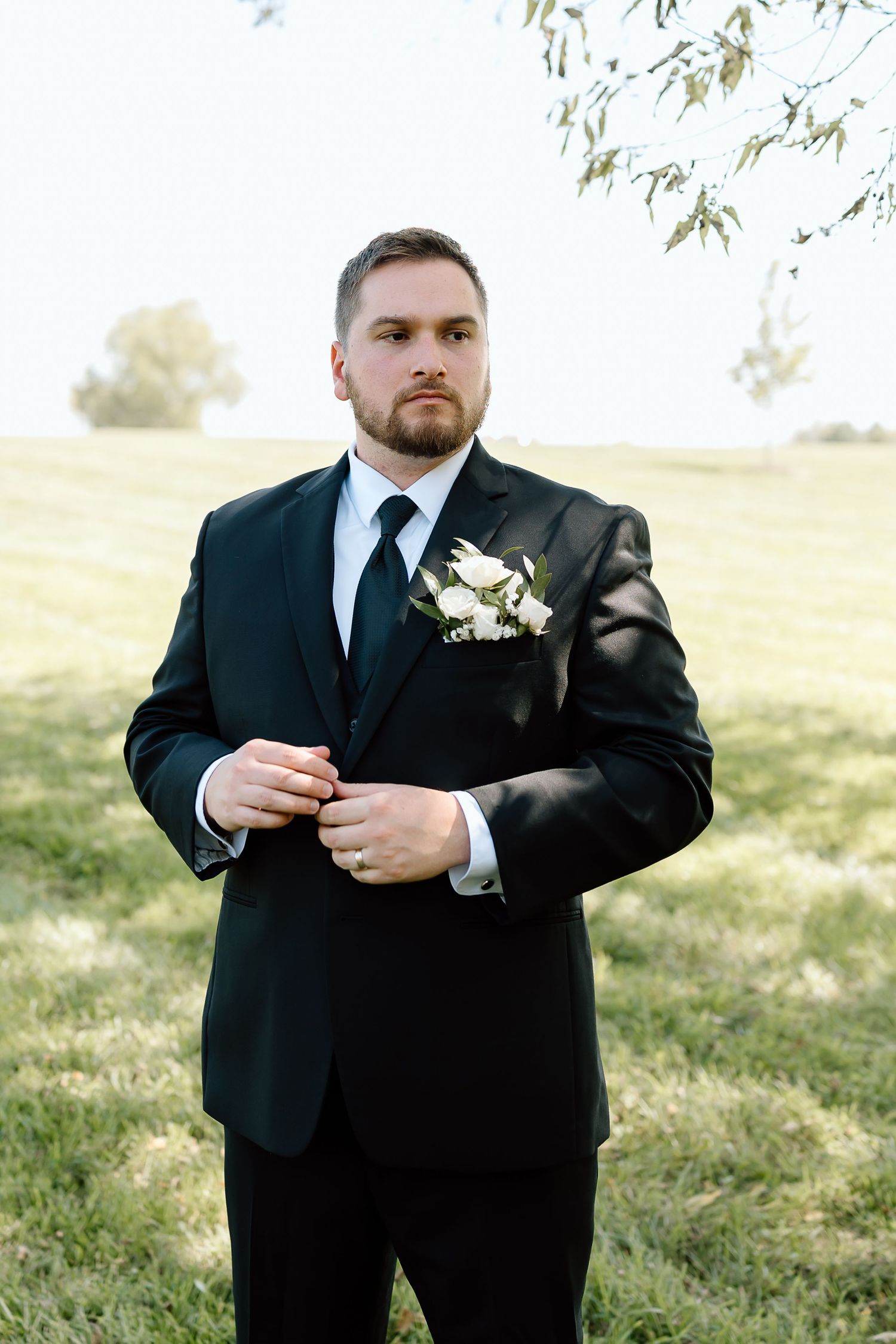 A groom in a black suit adjusts his tie while standing in a sunlit grassy field.
