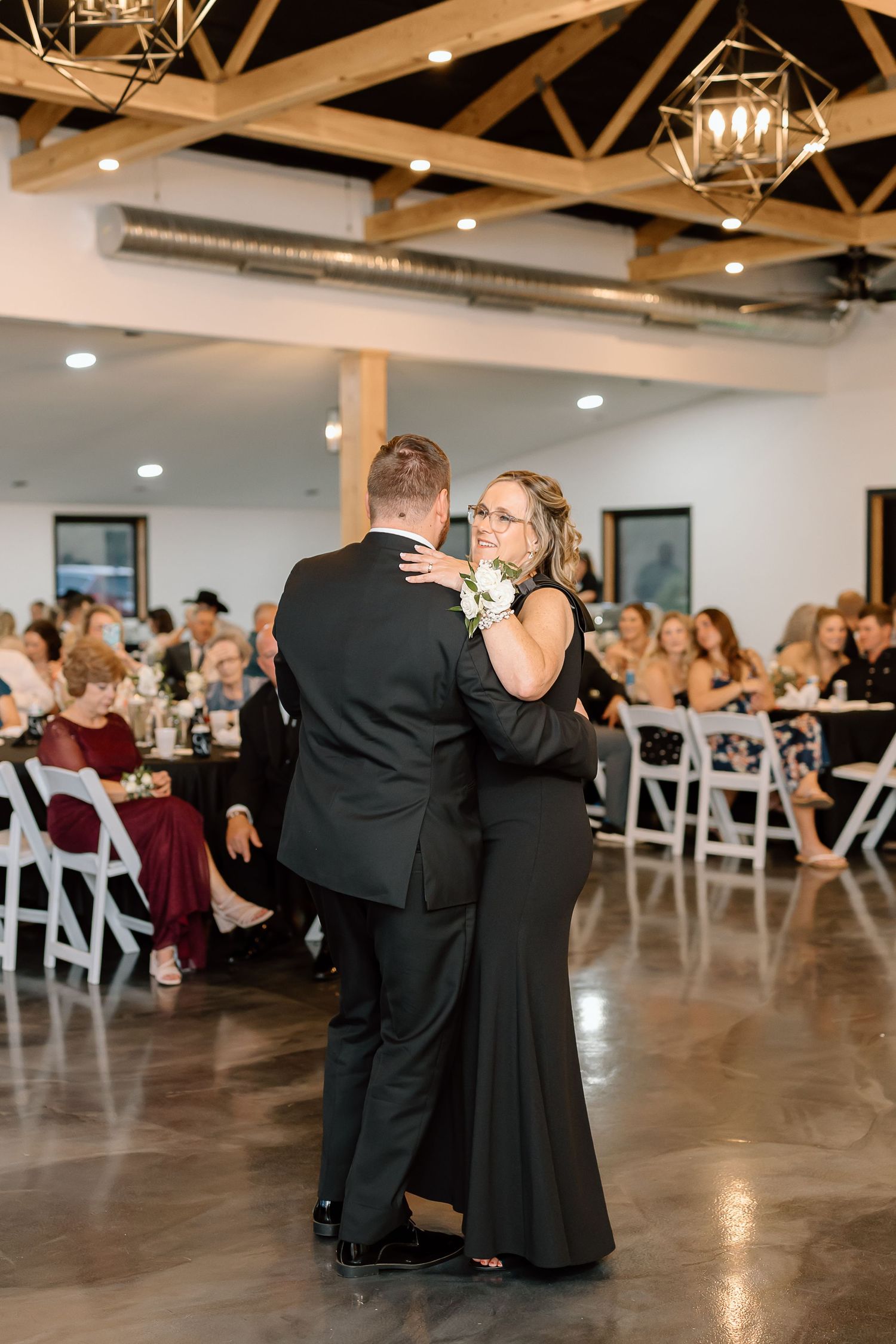 A couple shares a romantic dance during a wedding reception in an elegant venue with exposed wooden beams and white chairs.