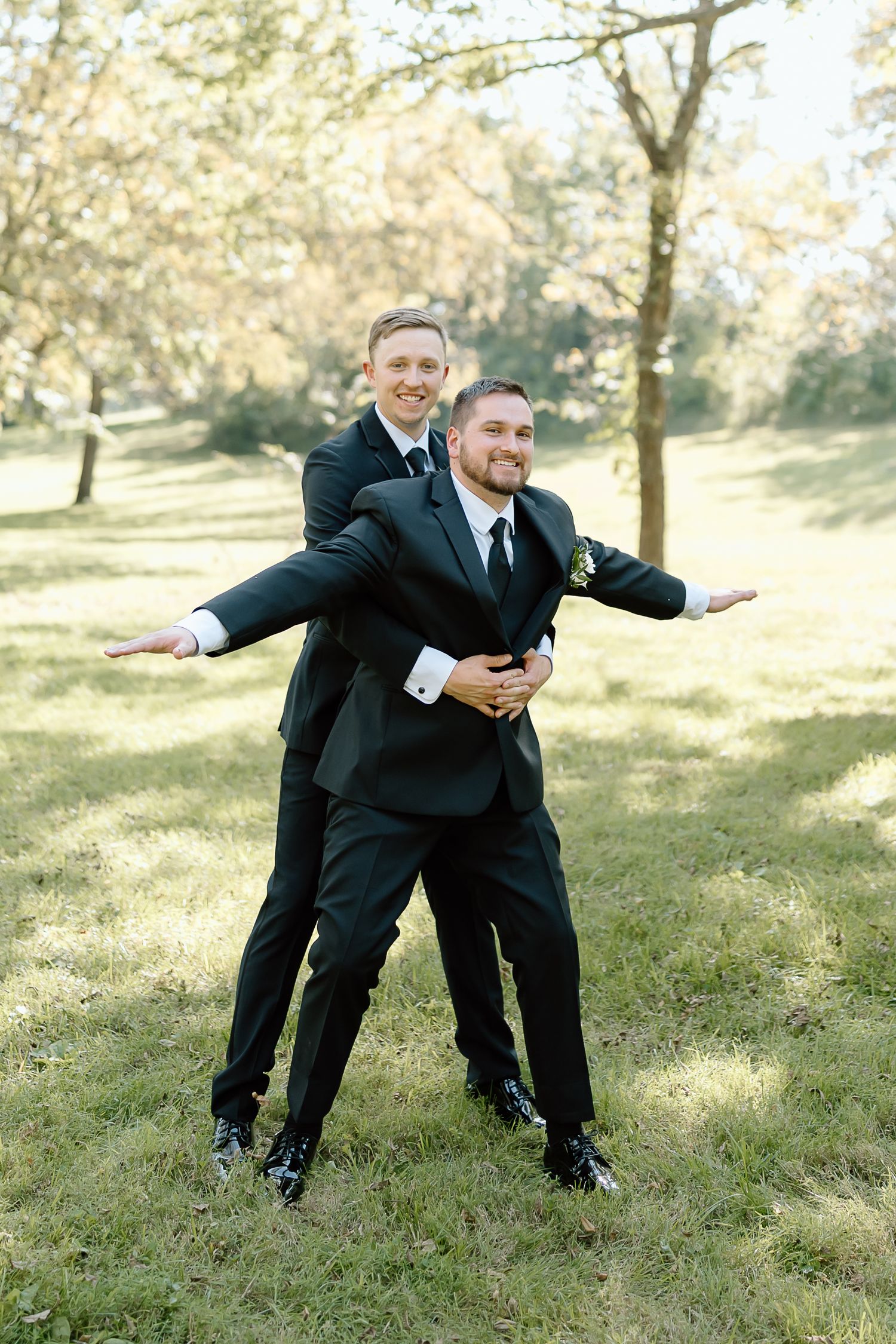 A sequence of playful poses showing someone in a black suit performing airplane motions on a grassy hillside.