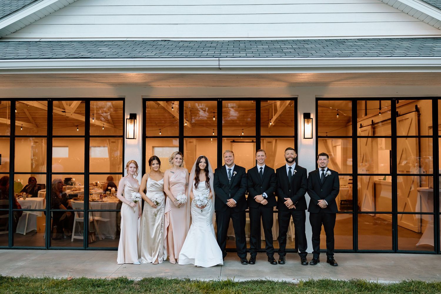Wedding party members sit in a row on a wooden deck in front of glass windows at an outdoor venue.