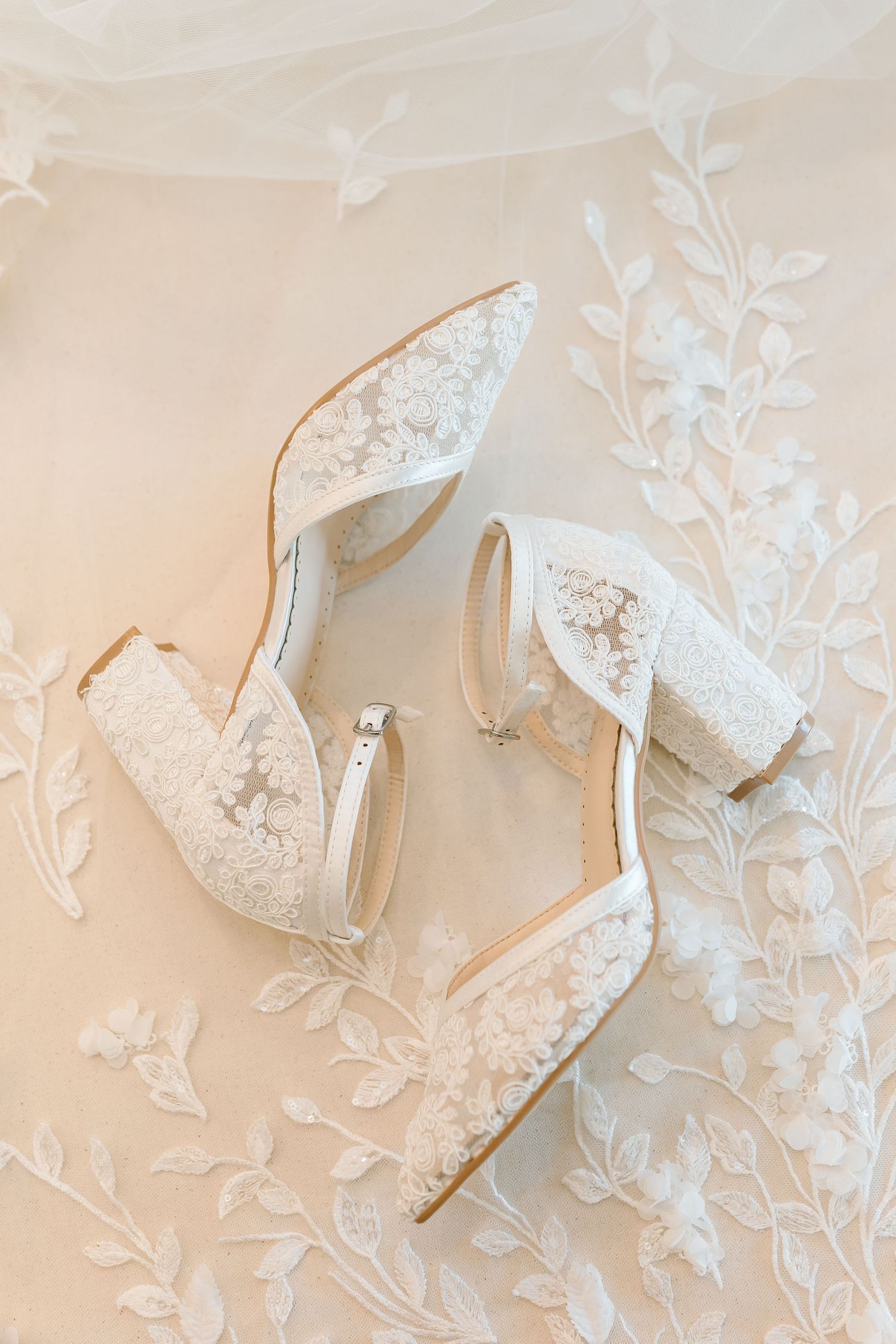 Elegant white lace bridal shoes displayed on delicate floral embroidered fabric in soft beige tones.