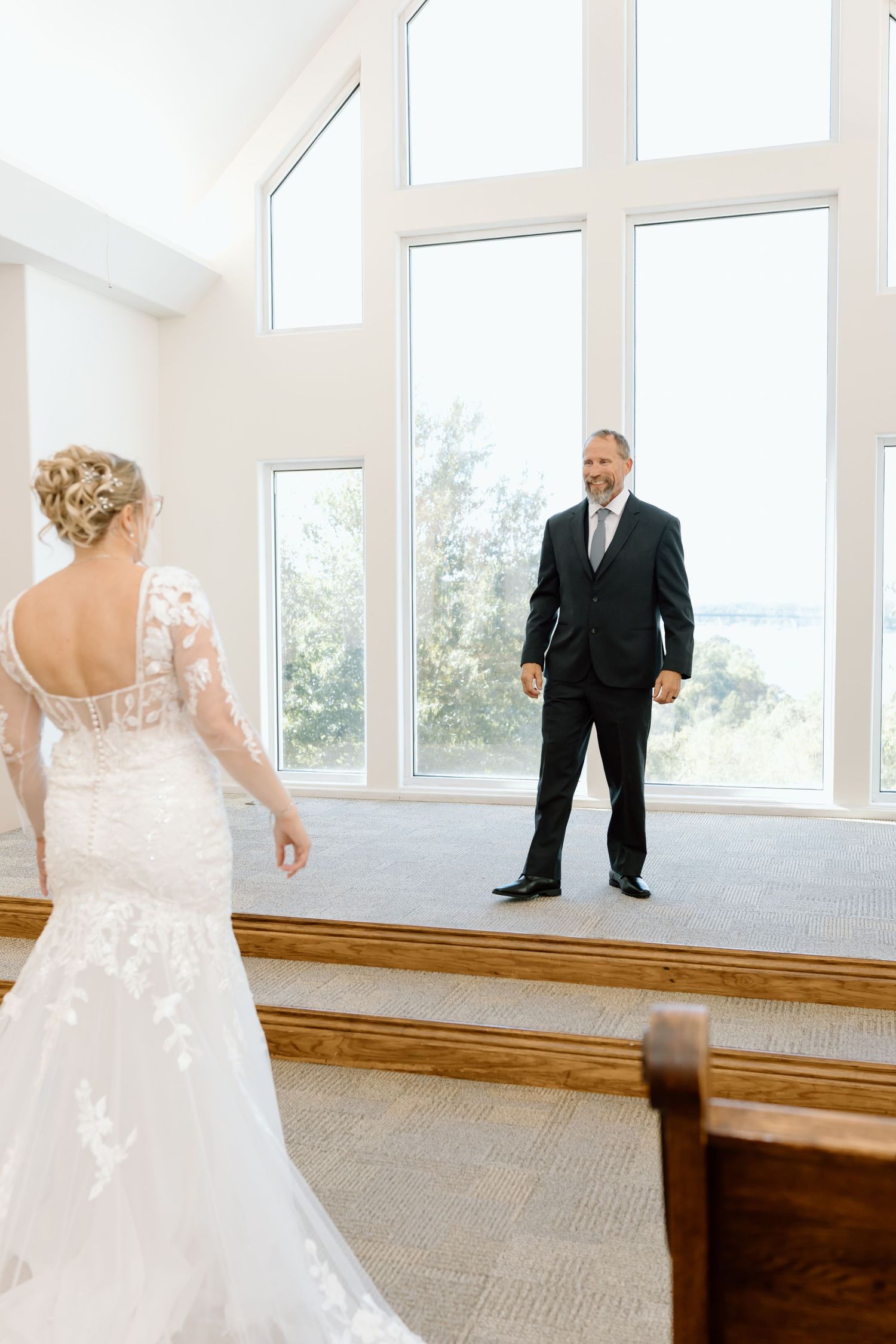 A heartwarming first look moment between a bride in a lace gown and a person in a dark suit in a bright sunlit room.