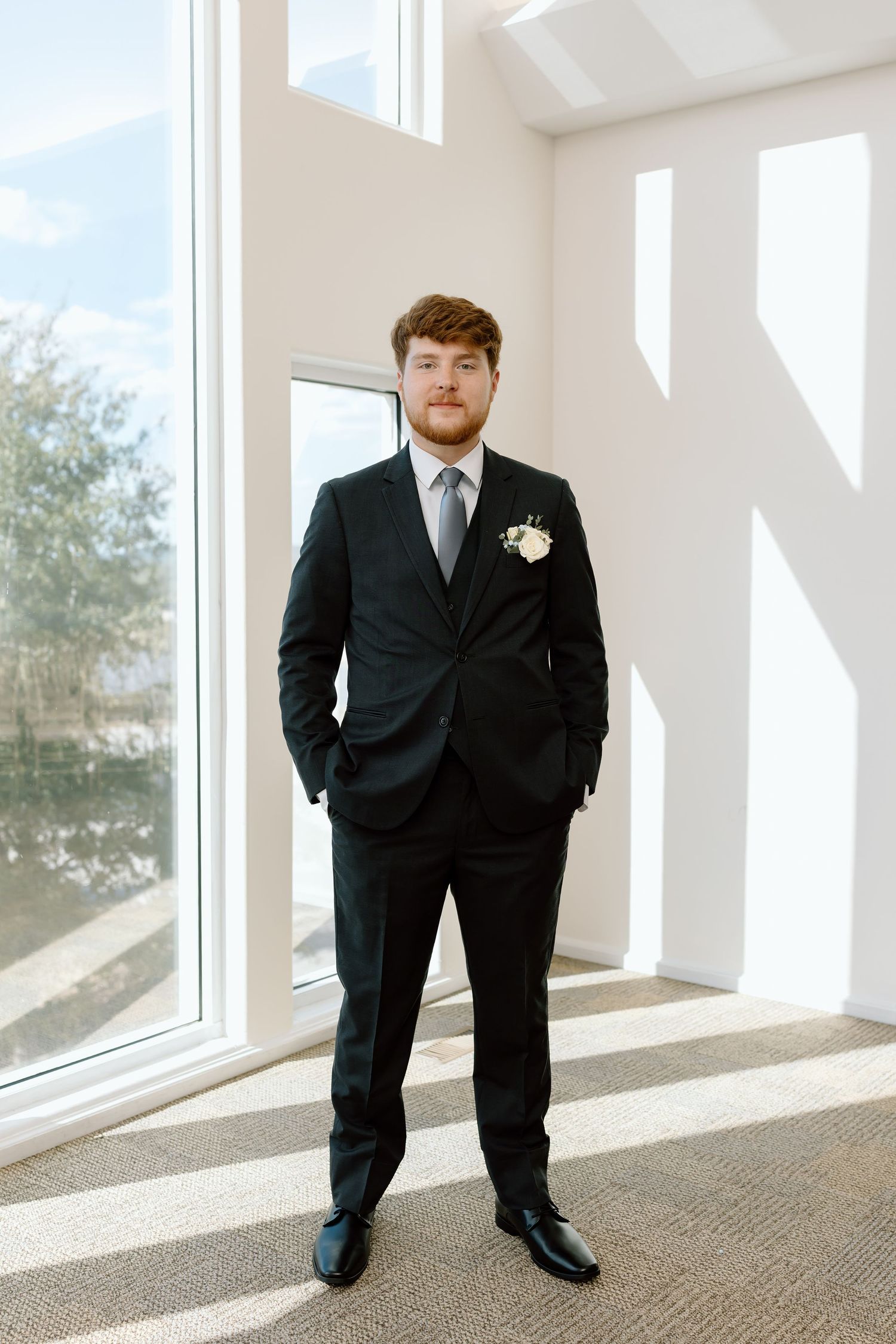 A groom in a classic black suit stands in a bright white room with large windows casting dramatic shadows.