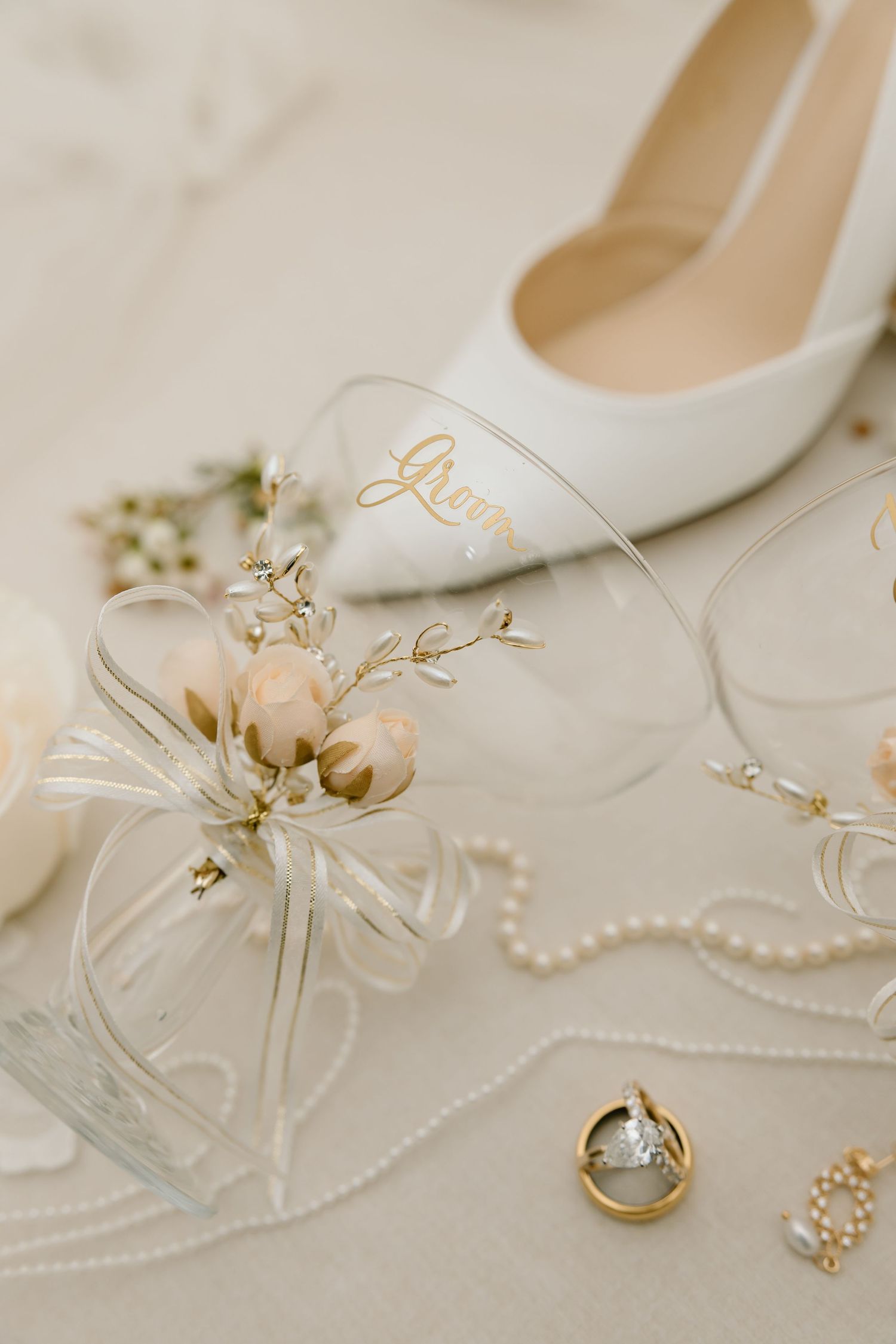 Elegant white bridal accessories including pearl necklace, wedding rings, and white heels arranged on light fabric.