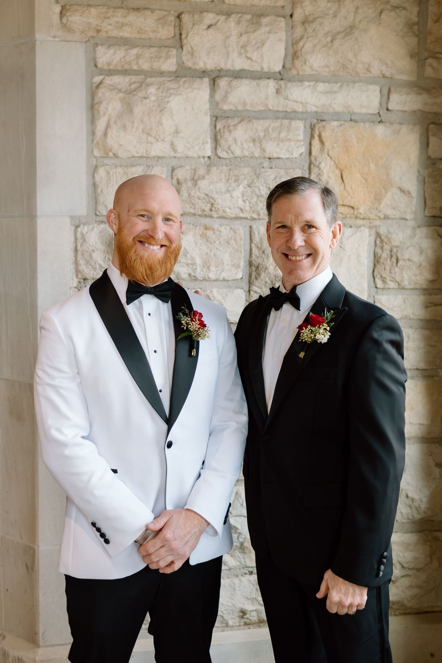 Two people in formal tuxedos with boutonnieres pose together against a stone wall backdrop.