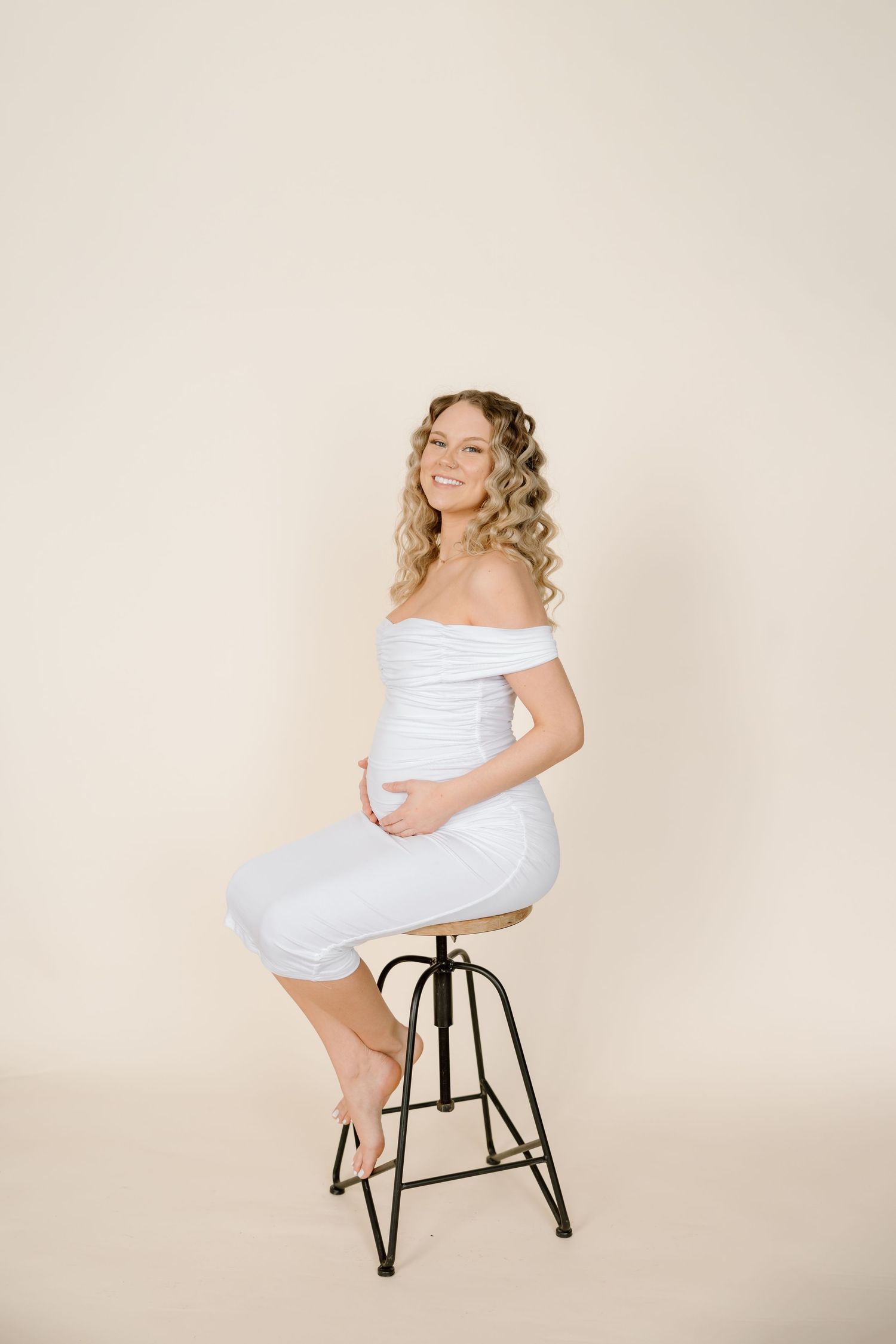 A pregnant woman in a white off-shoulder dress sits elegantly on a black metal stool against a beige backdrop.