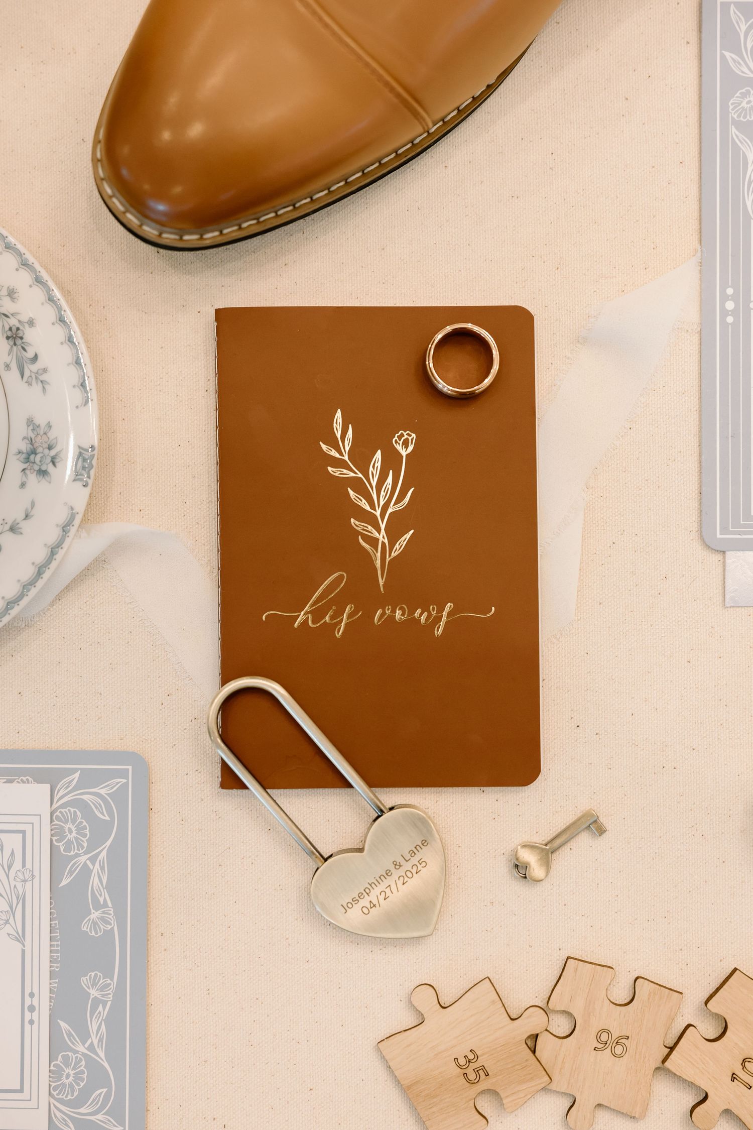 Flatlay of brown leather wedding vow book with calligraphy, heart lock, ring, and puzzle pieces on beige background.