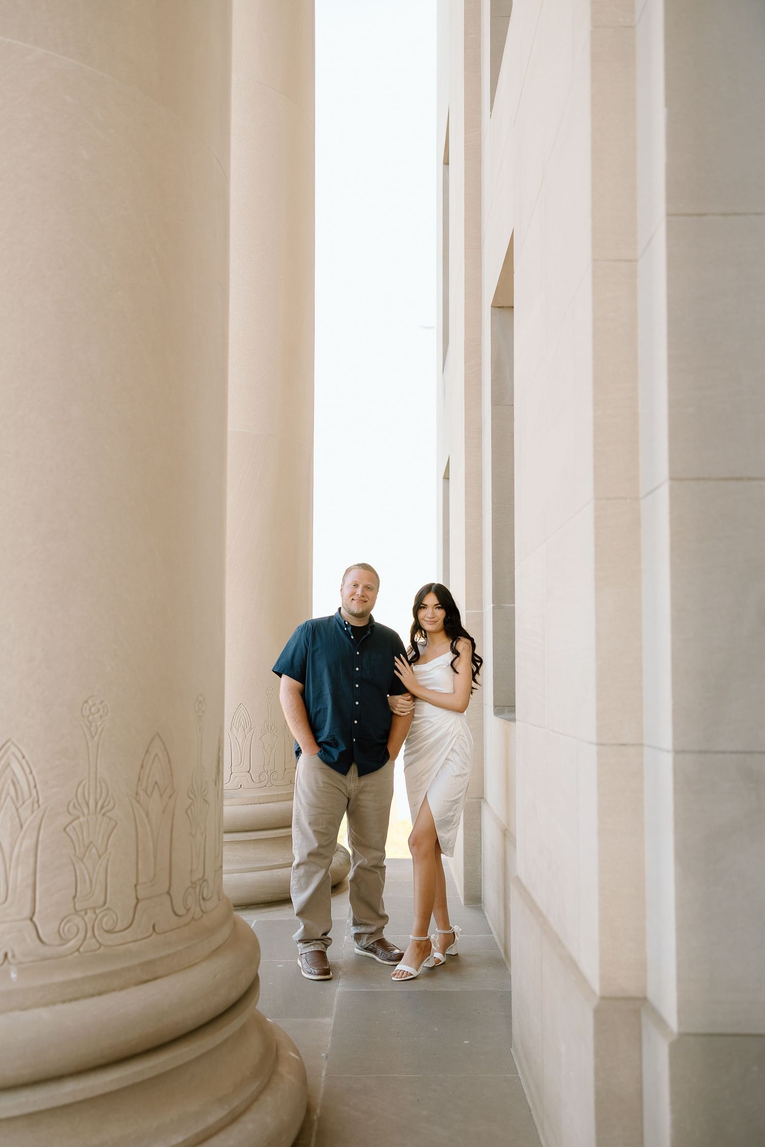 A couple poses playfully between white marble columns in an elegant architectural setting.