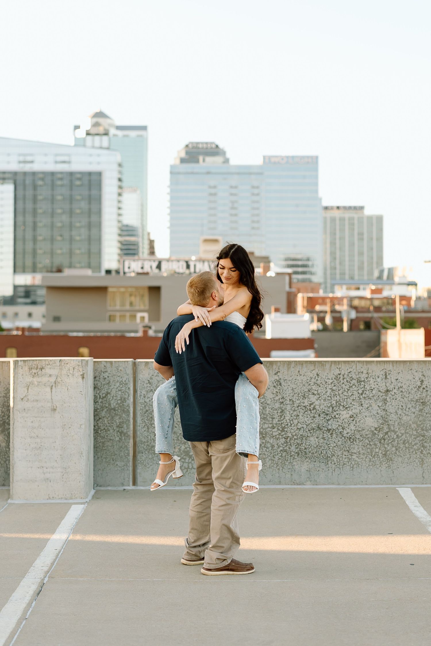 Two people share a romantic moment on a rooftop parking deck with a city skyline backdrop at sunset.