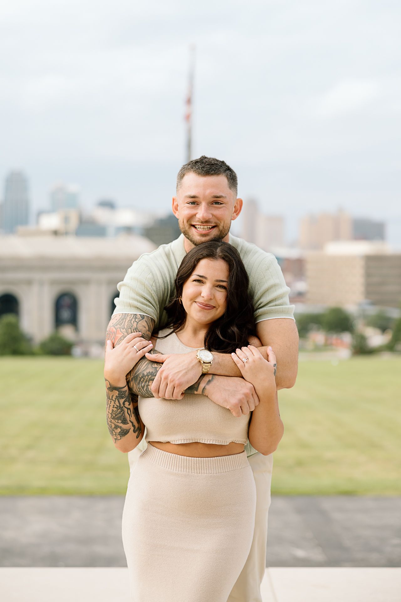 A couple embraces in a romantic moment with a city skyline backdrop on a grassy hill.