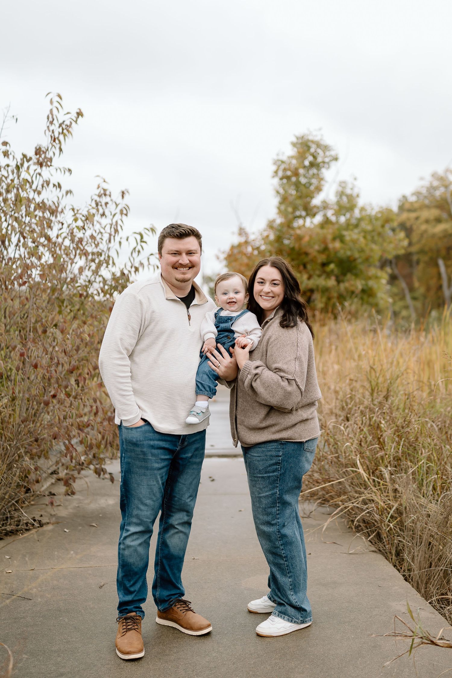 Family in matching denim jeans and light tops pose together on a path surrounded by autumn grasses and plants.