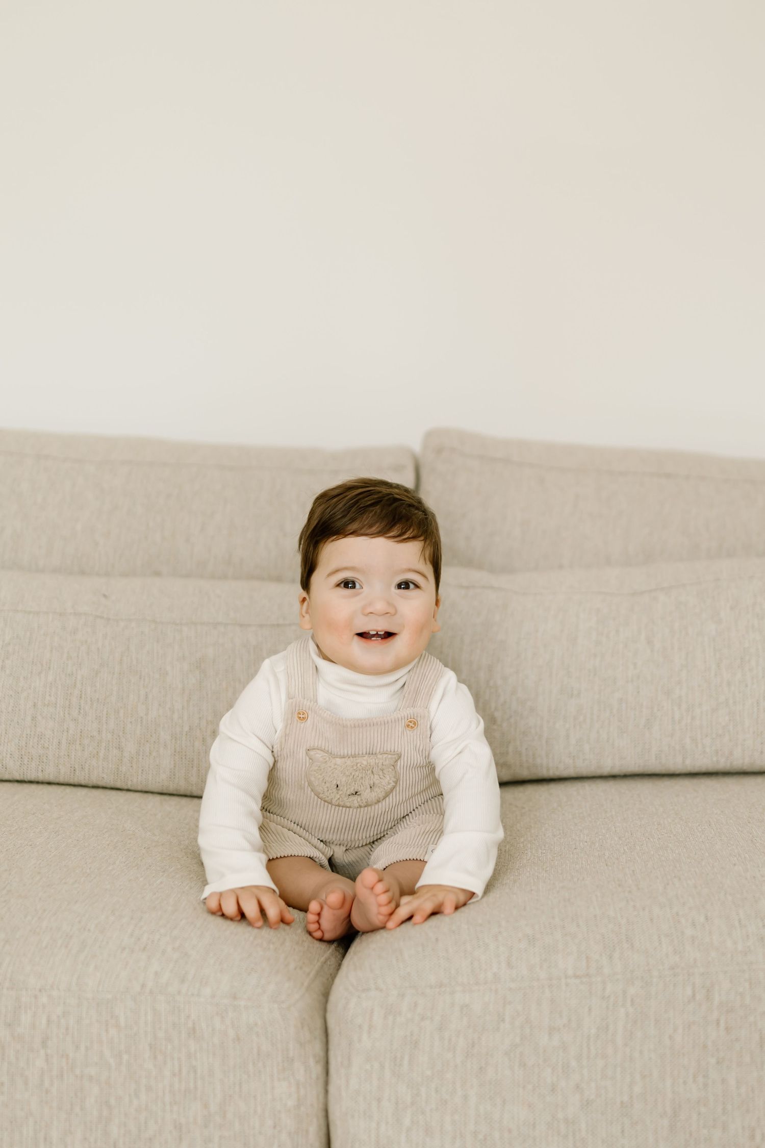 Small child wearing beige overalls and white shirt sits happily on a light gray couch.