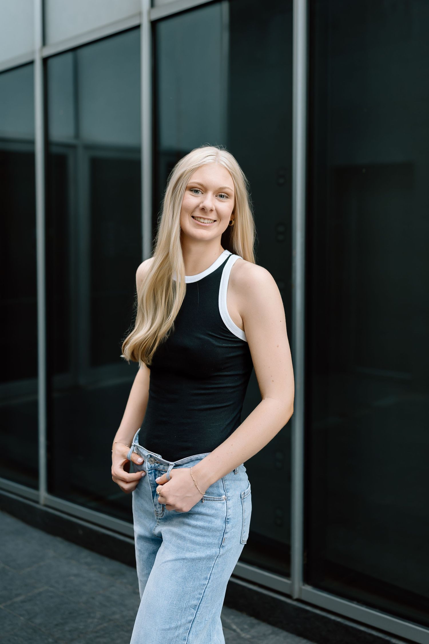 A person in a black tank top and light jeans poses casually against a glass window wall.