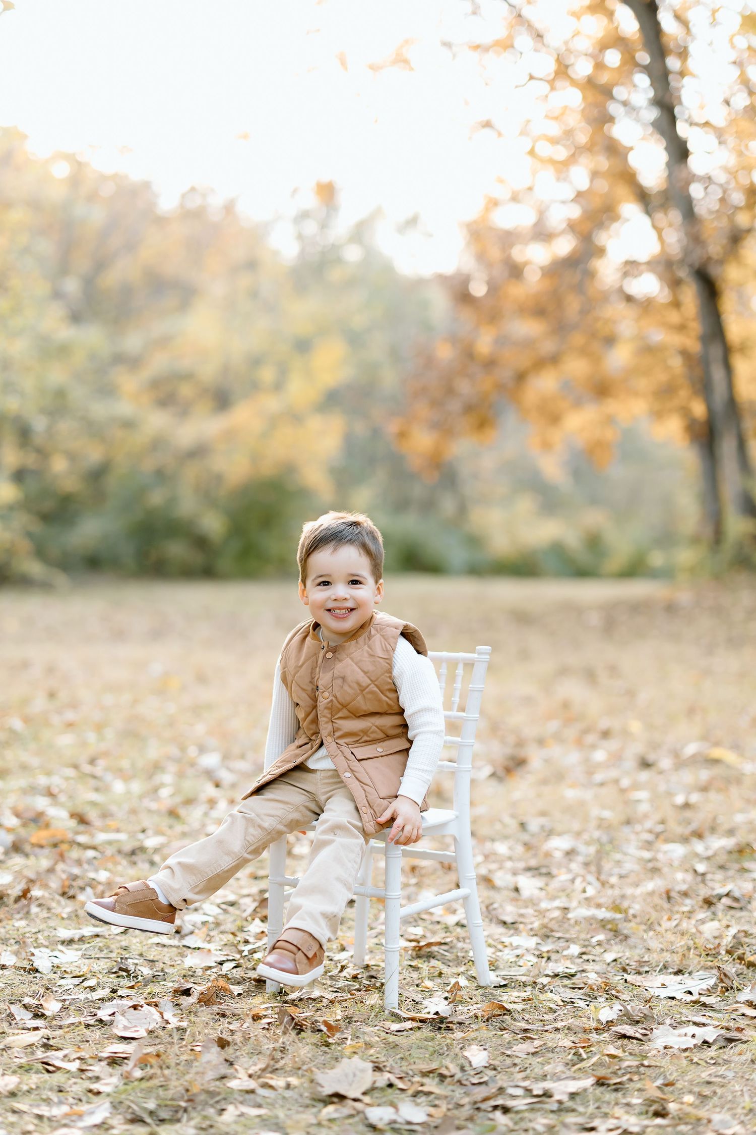 A child in brown clothing plays in autumn leaves near a white chair in a natural outdoor setting.