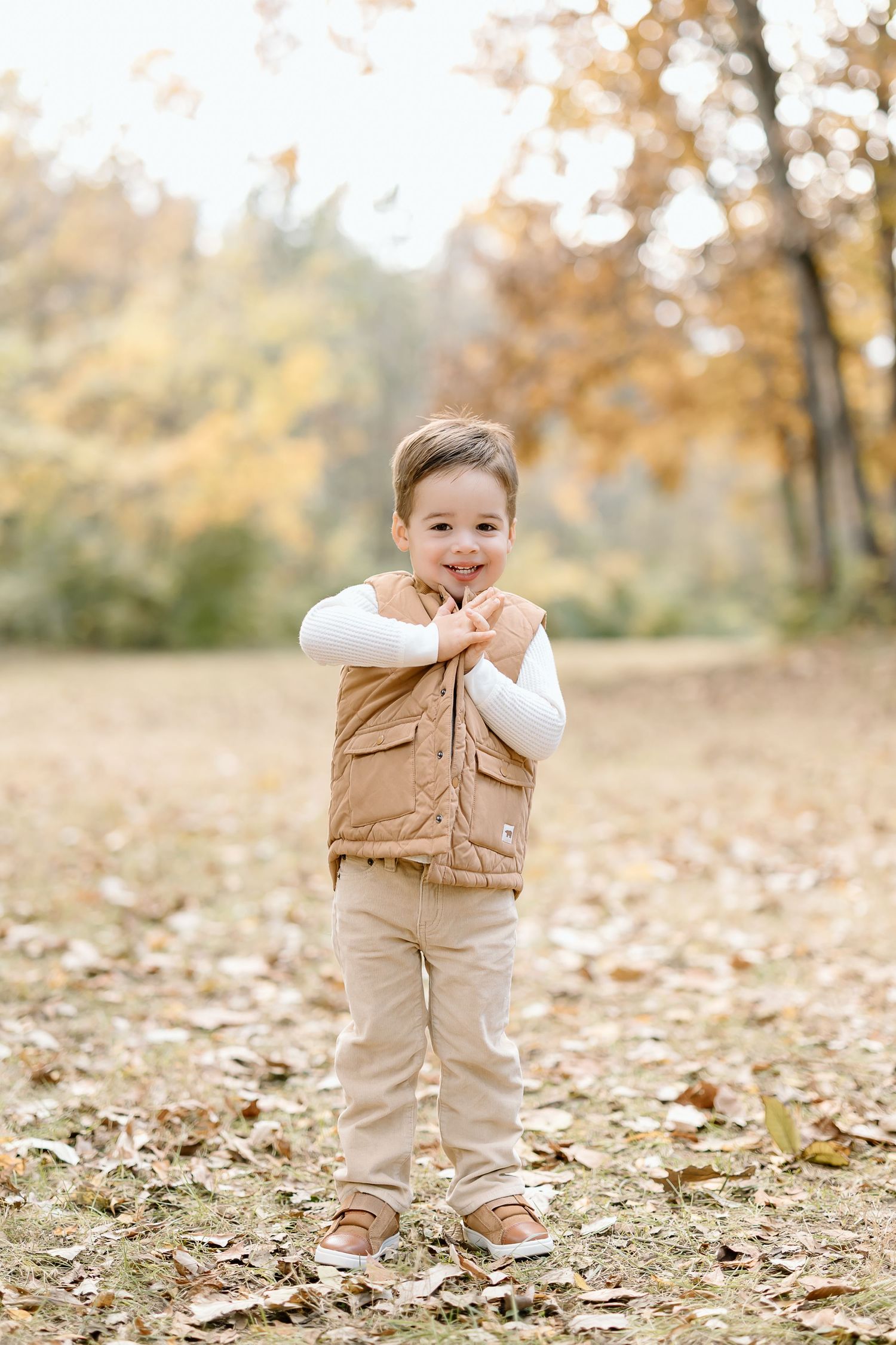 A child in a tan vest and white shirt plays among fallen autumn leaves in a sunlit wooded area.