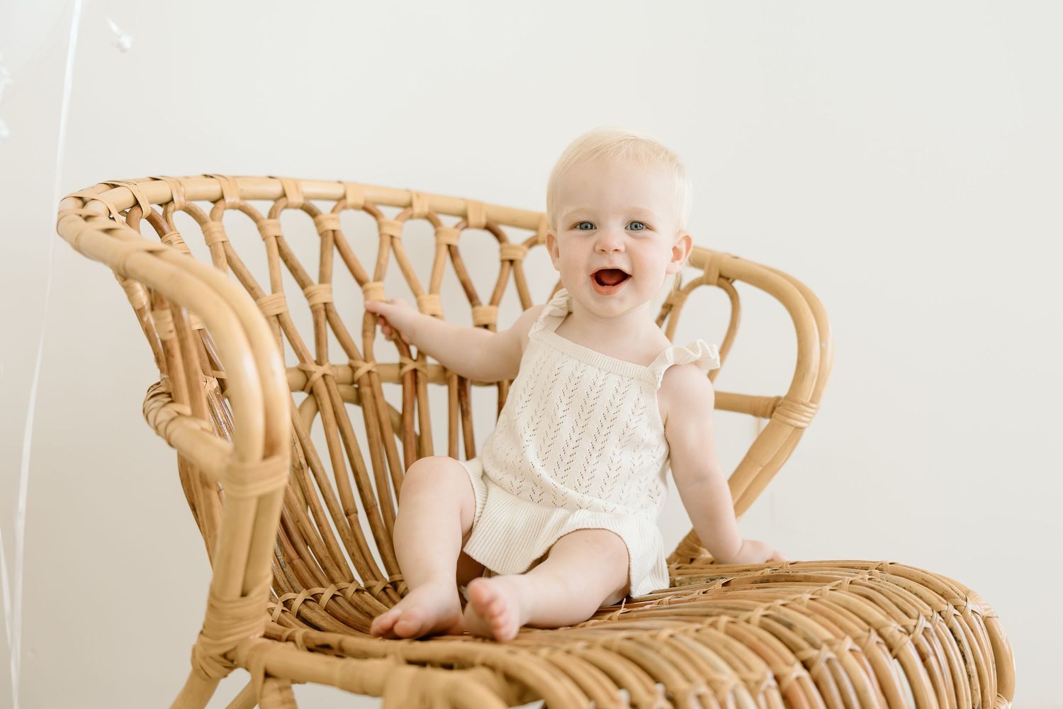 A laughing baby wearing white sits on a vintage rattan peacock chair against a light colored wall.