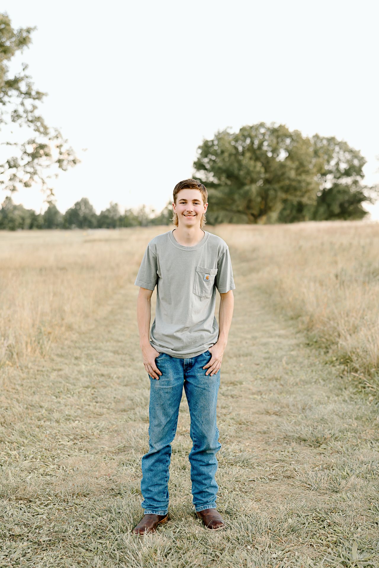 A person stands on a dirt path in a golden field wearing a gray t-shirt and blue jeans on a sunny day.