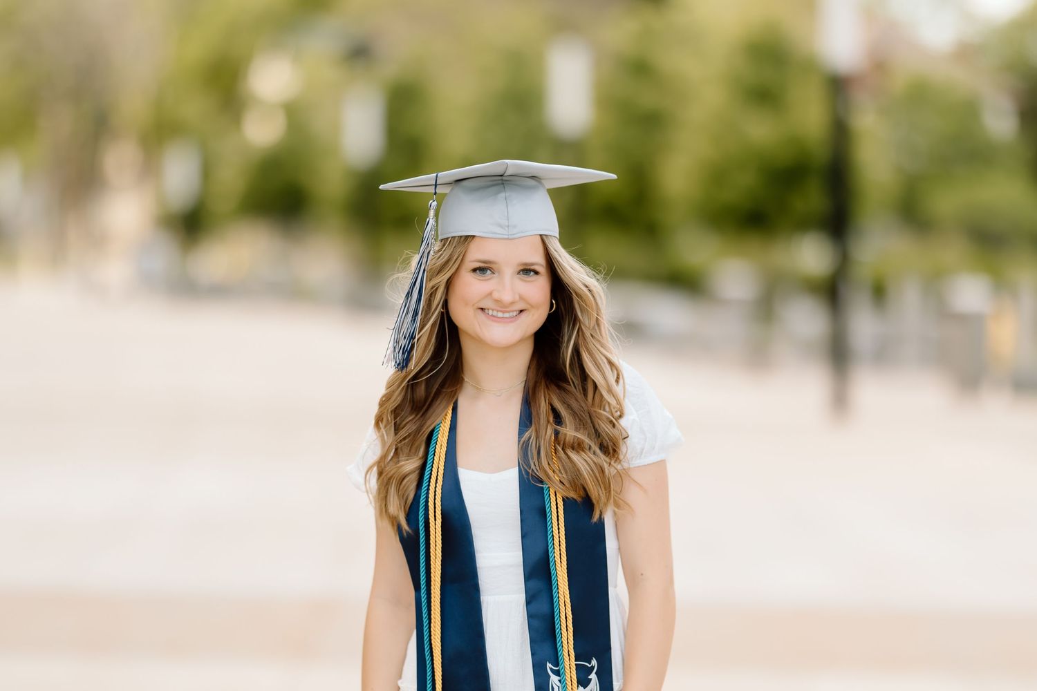 Graduate in blue cap and gown poses for graduation photos outdoors on a sunny day.