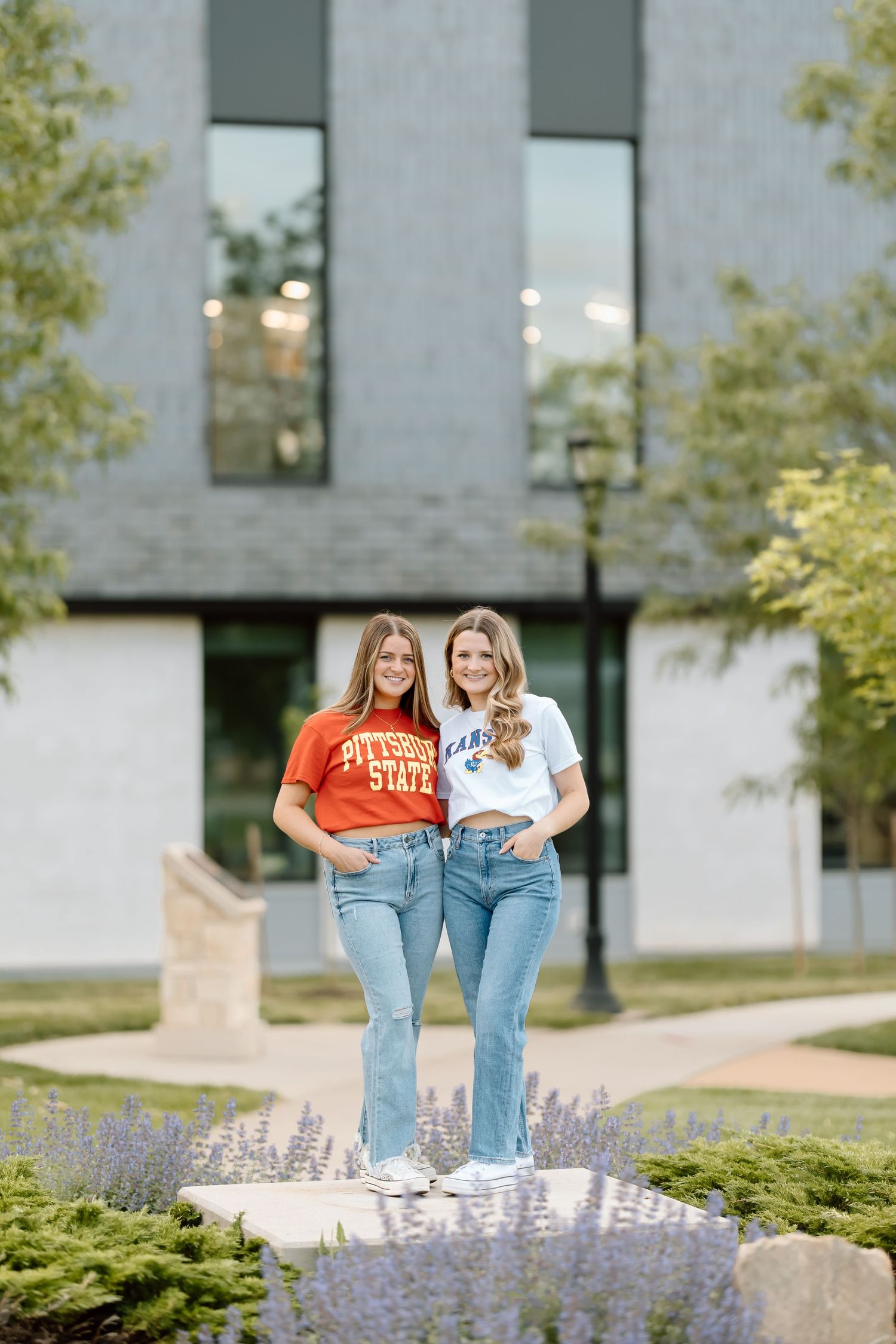 Two people in casual outfits standing together on a landscaped path with lavender flowers and modern building in background.